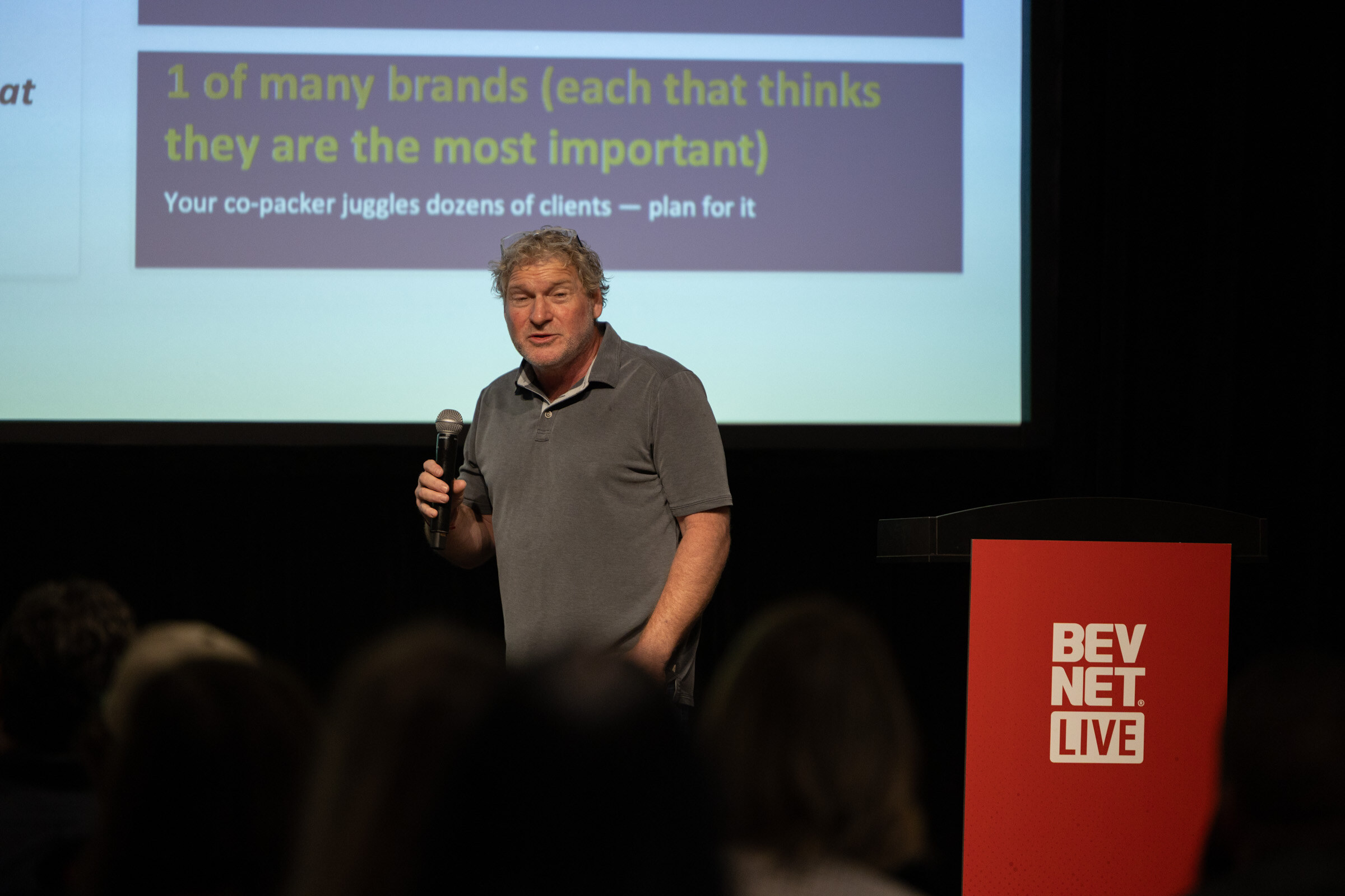 Man in a grey polo speaking into a microphone in front of a slide about co-packers juggling dozens of clients.