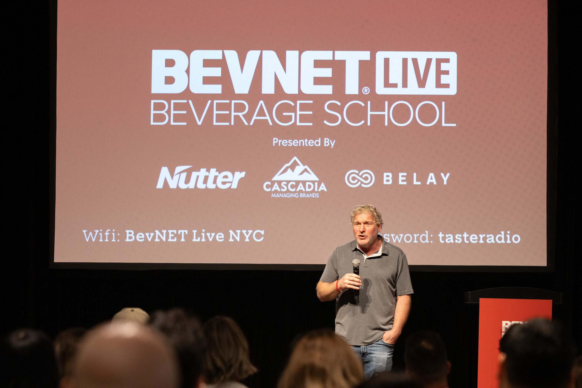 Man in a grey polo with a microphone standing in front of the BevNET Live Beverage School sponsor slide.