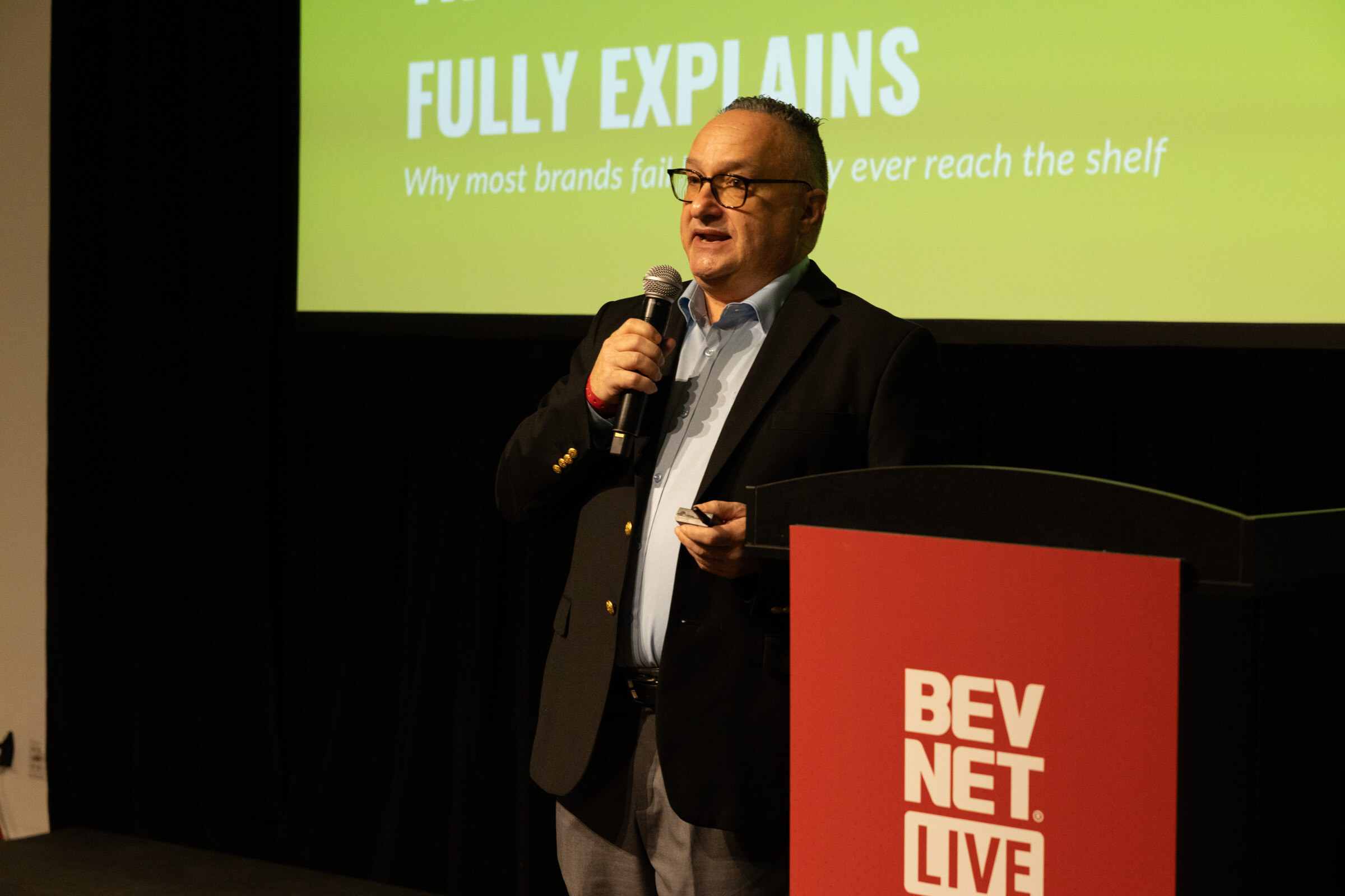 Man in a dark blazer speaking into a microphone at a red BevNET Live podium under a green slide.