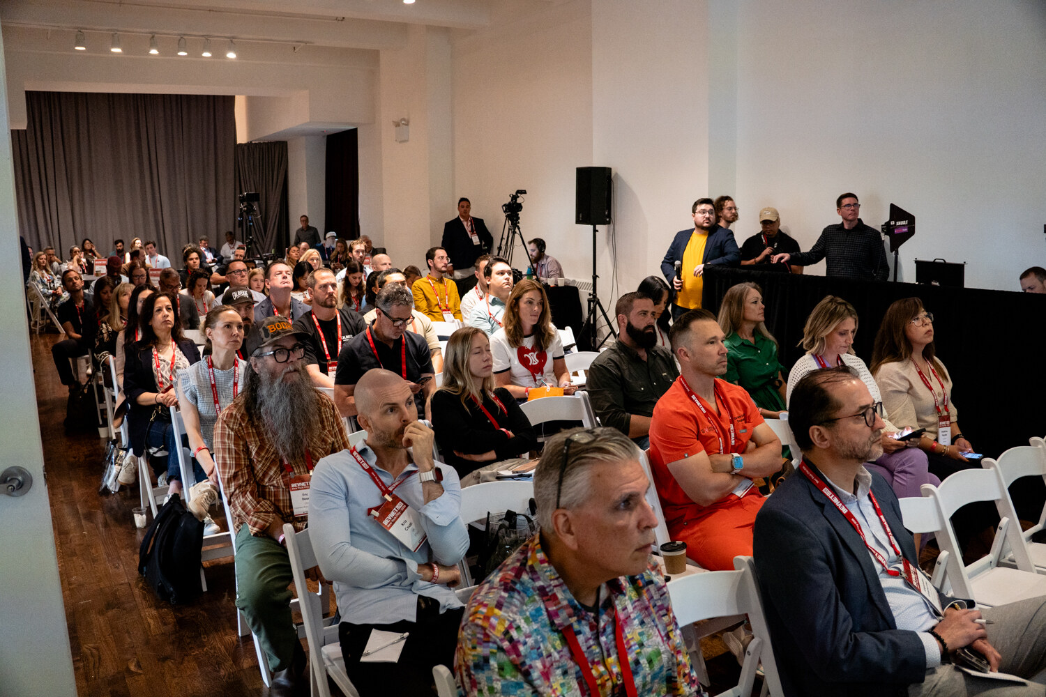 Wide view of a crowded room of seated attendees with lanyards, cameras on tripods at the back.