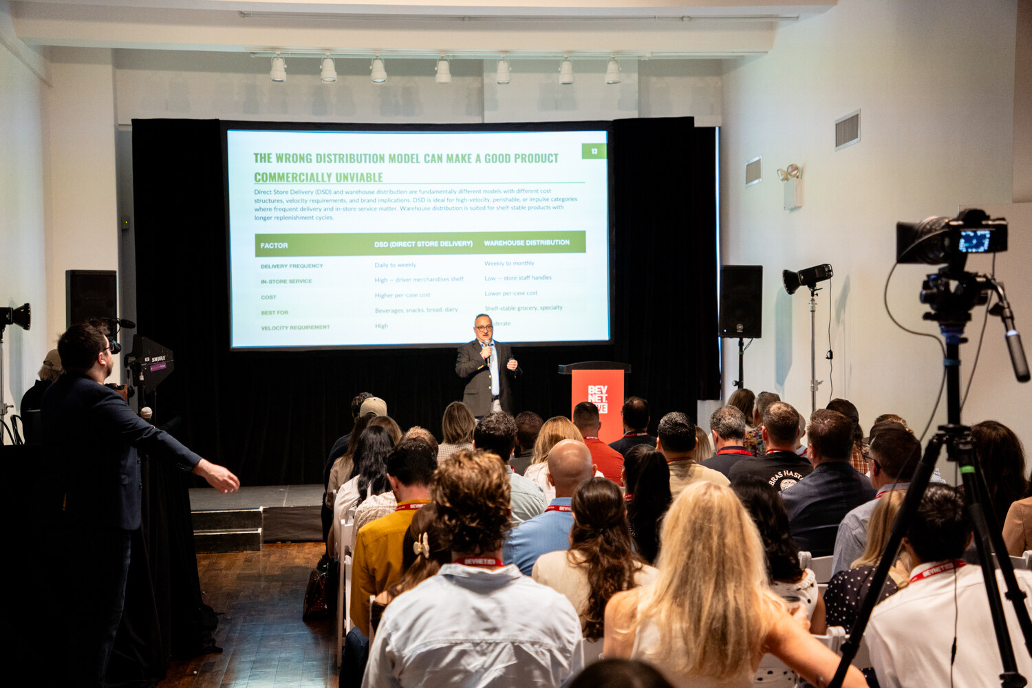Speaker at a red BevNET Live podium beneath a distribution slide, seen past attendees and a camera on a tripod.