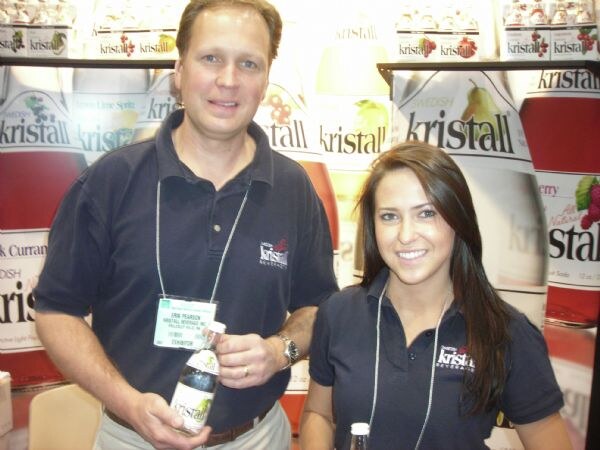 Erin Pearson of Kristall, in a navy polo and exhibitor badge, holds a Kristall bottle beside a woman with long brown hair in a matching polo, in front of Kristall bottle graphics.