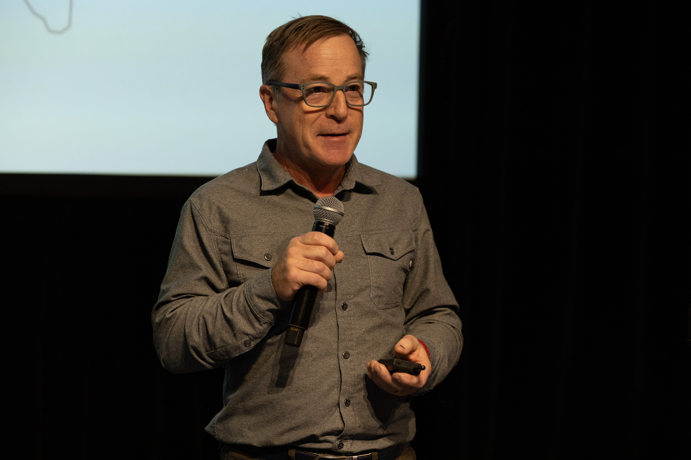 Man in glasses and a grey shirt speaking into a microphone while holding a slide clicker.
