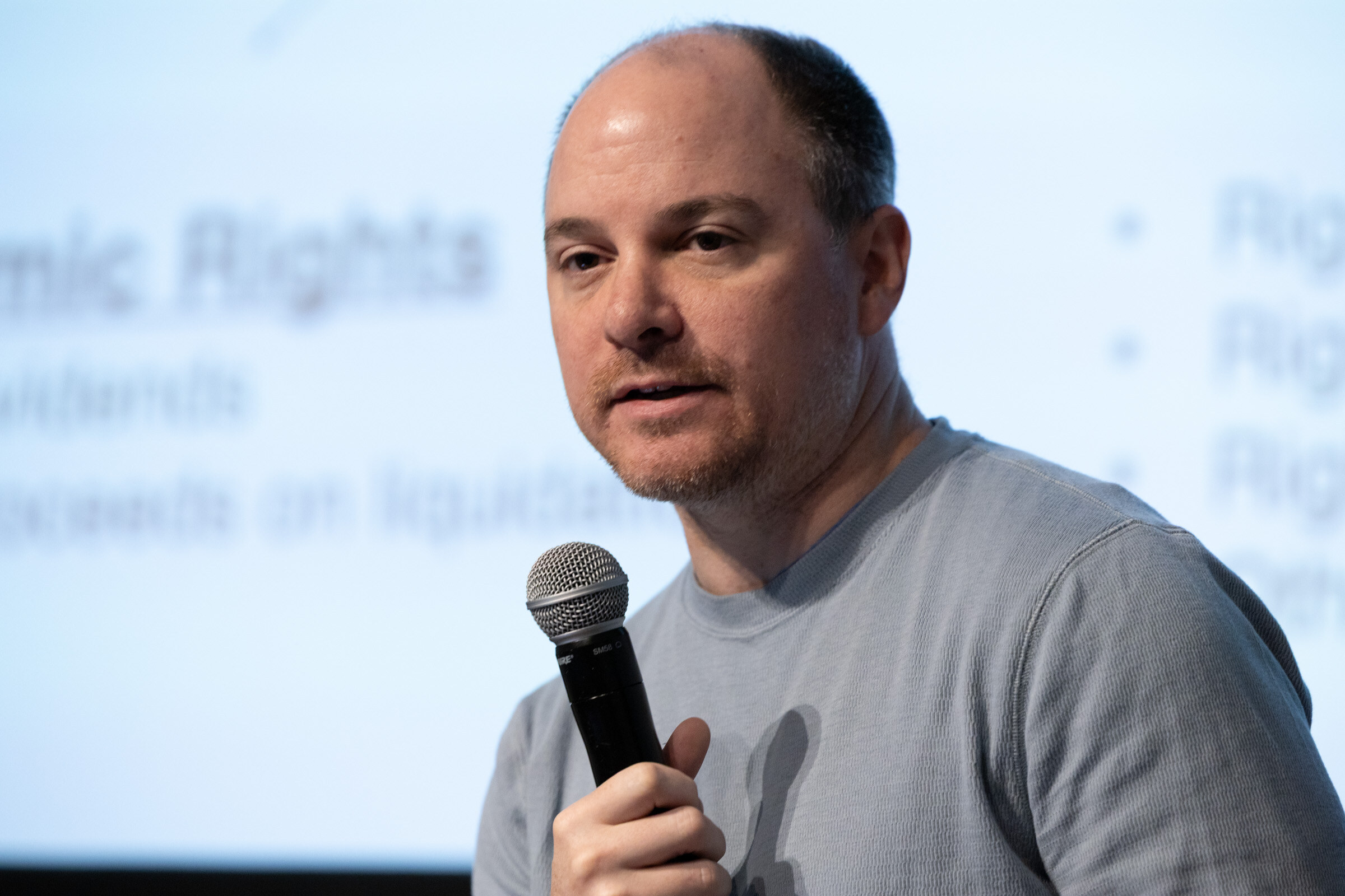 Man in a grey sweater speaking into a microphone in front of a slide about economic rights.