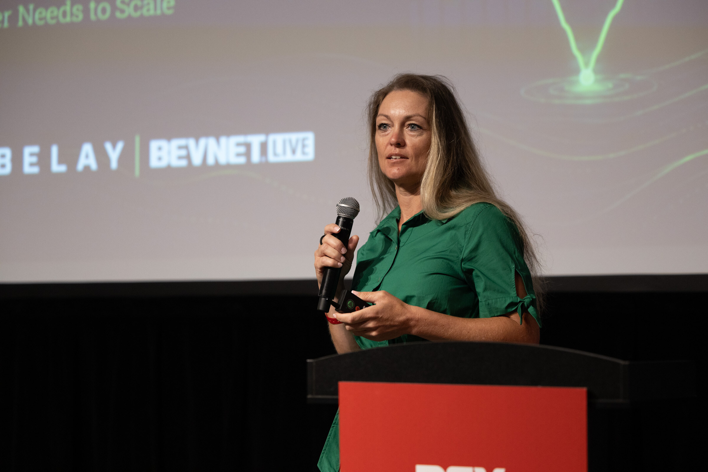 Woman in a green dress speaking at a podium in front of a Belay and BevNET Live slide.