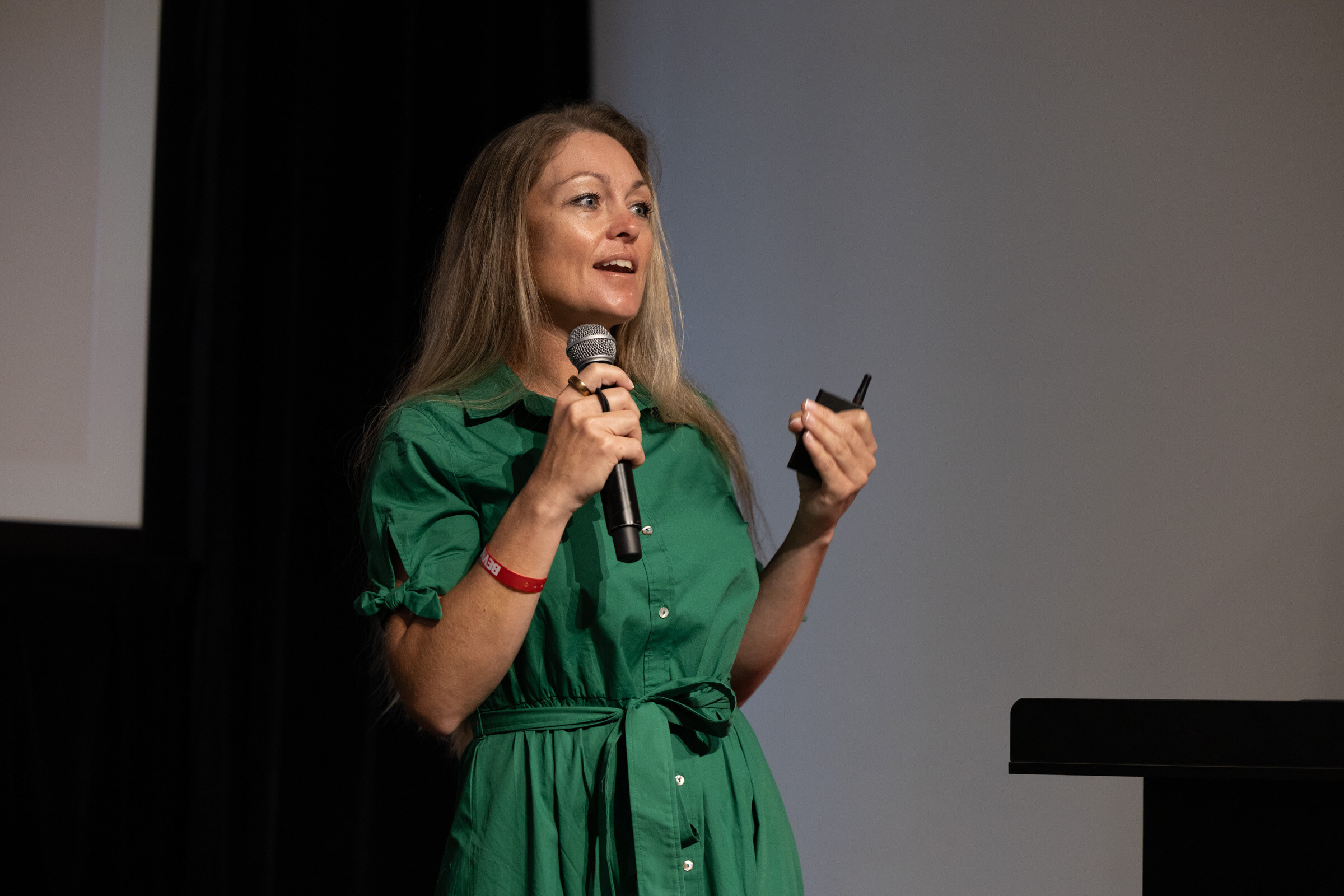 Woman in a green dress speaking into a microphone and holding a clicker on a dark stage.