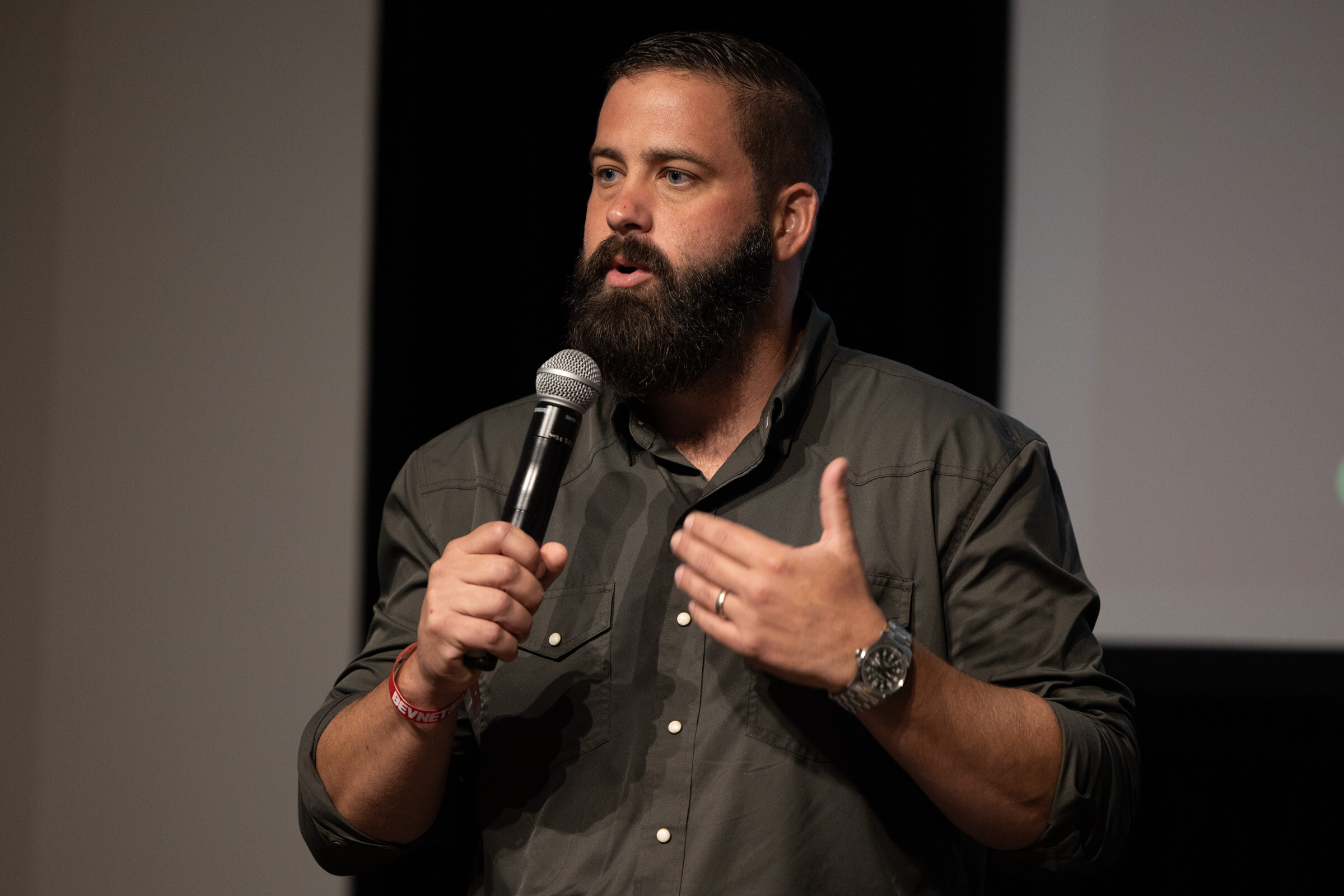 Bearded man in an olive shirt speaking into a microphone and gesturing.