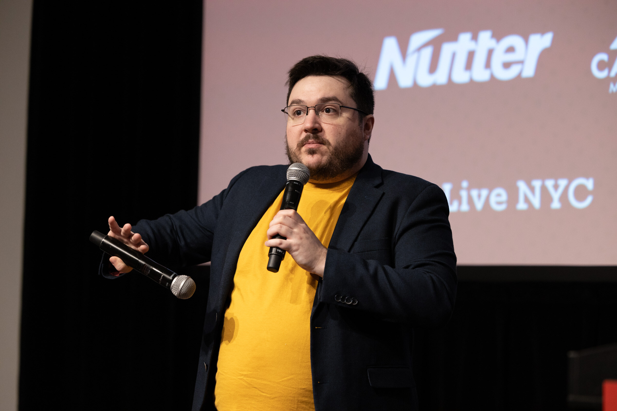 Man in a navy blazer and yellow T-shirt holding two microphones, a Nutter slide behind him.