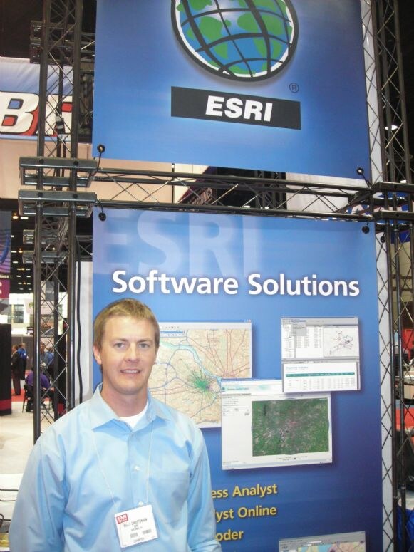 A smiling man in a light blue shirt and badge stands in front of an ESRI Software Solutions display showing map and software screenshots on a metal truss frame.
