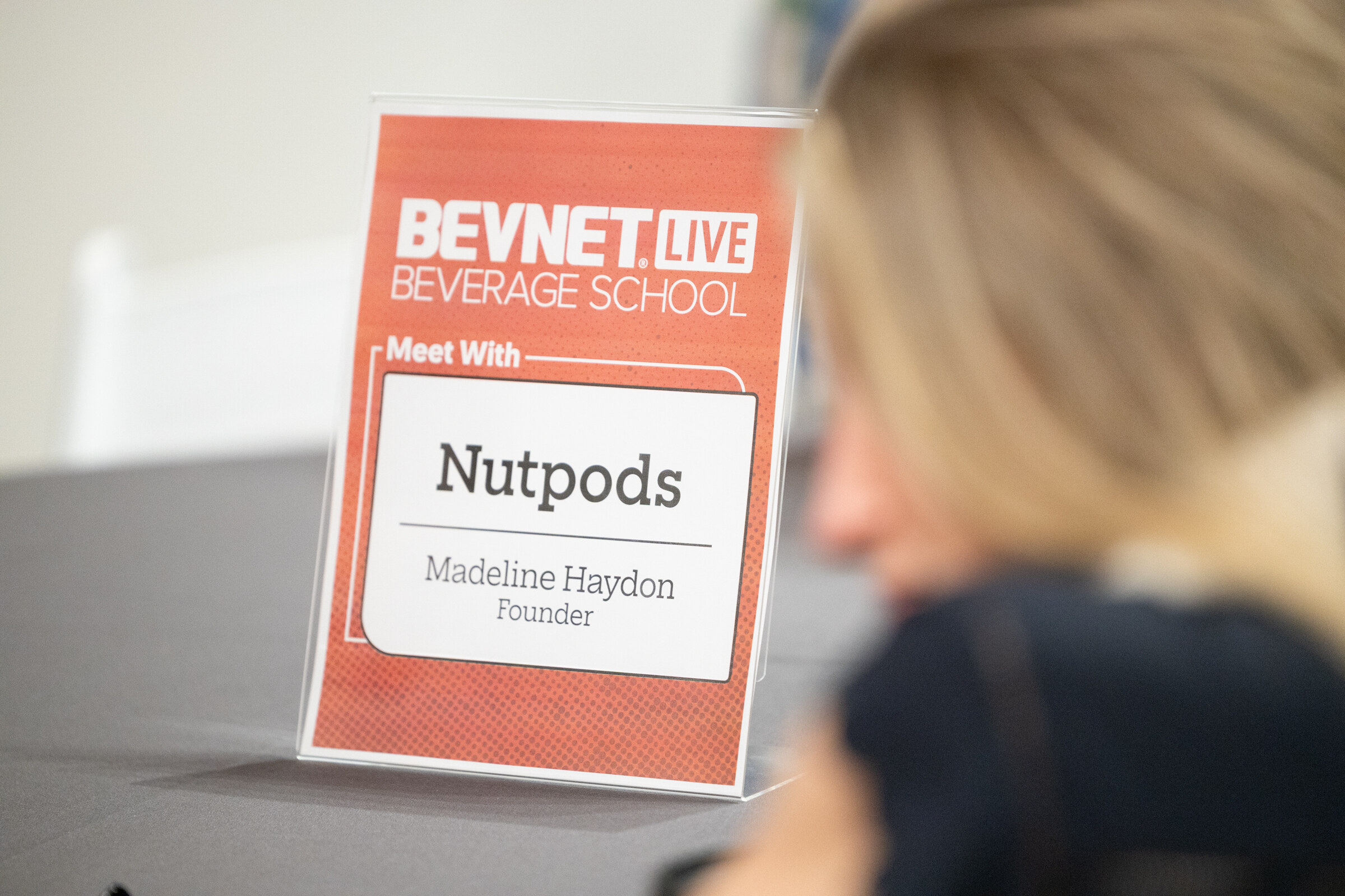 Red Beverage School “Meet With Nutpods” table card on a dark tablecloth, an attendee blurred in the foreground.
