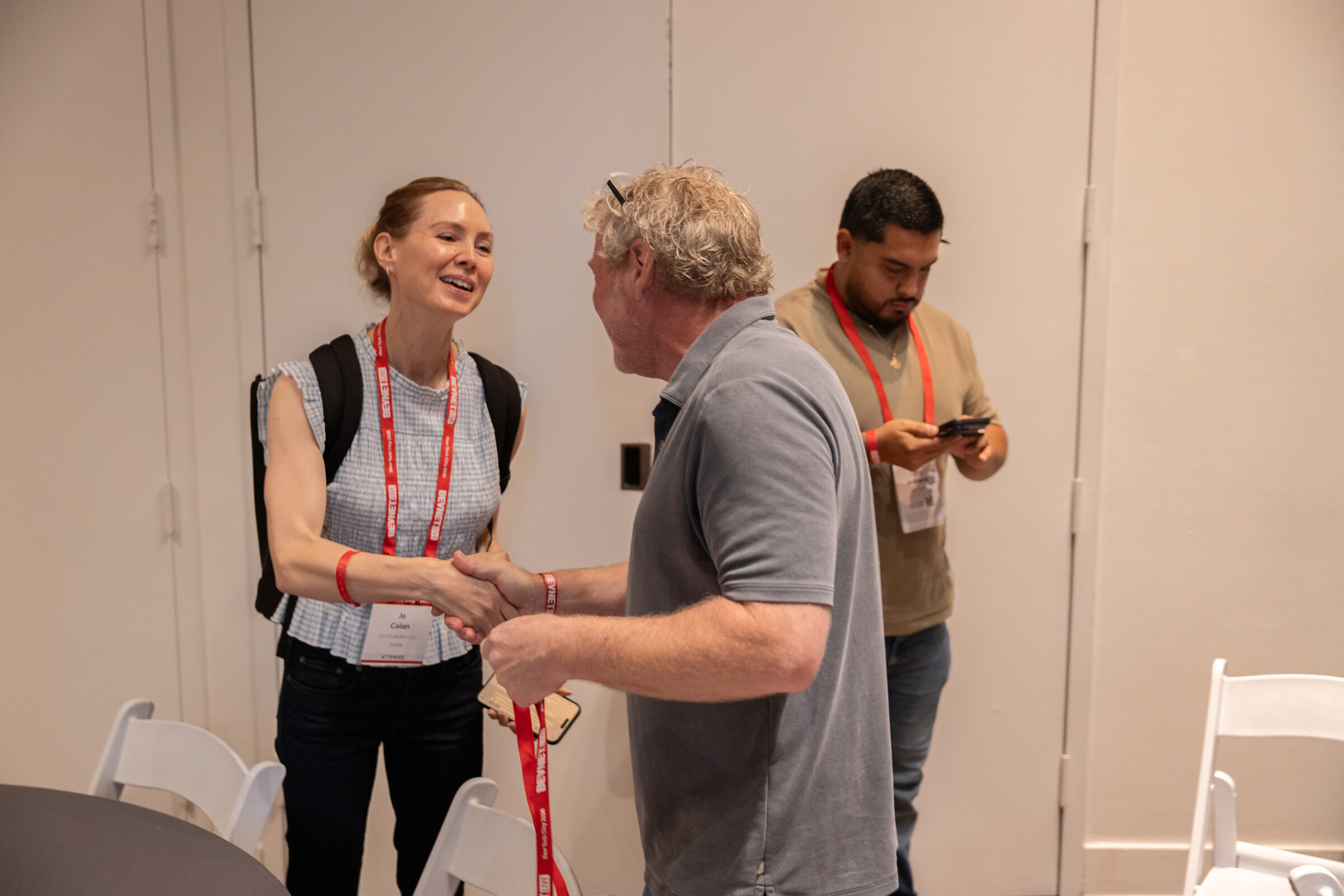 Smiling woman with a backpack shaking hands with a man in a grey polo while another attendee checks his phone.