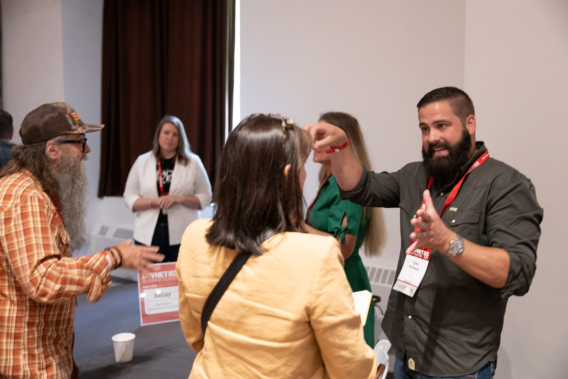 Bearded man gesturing animatedly to a group of attendees beside a Belay table card.