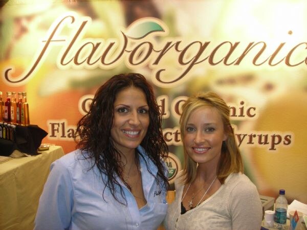 Two smiling women pose close together in front of a Flavorganics banner for organic flavor syrups, with syrup bottles on a table at left.