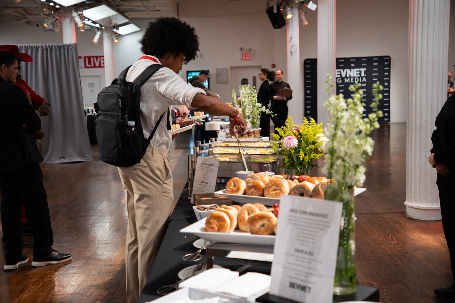 Attendee with a backpack reaching across a breakfast table of bagels, flowers and a New York Breakfast menu card.