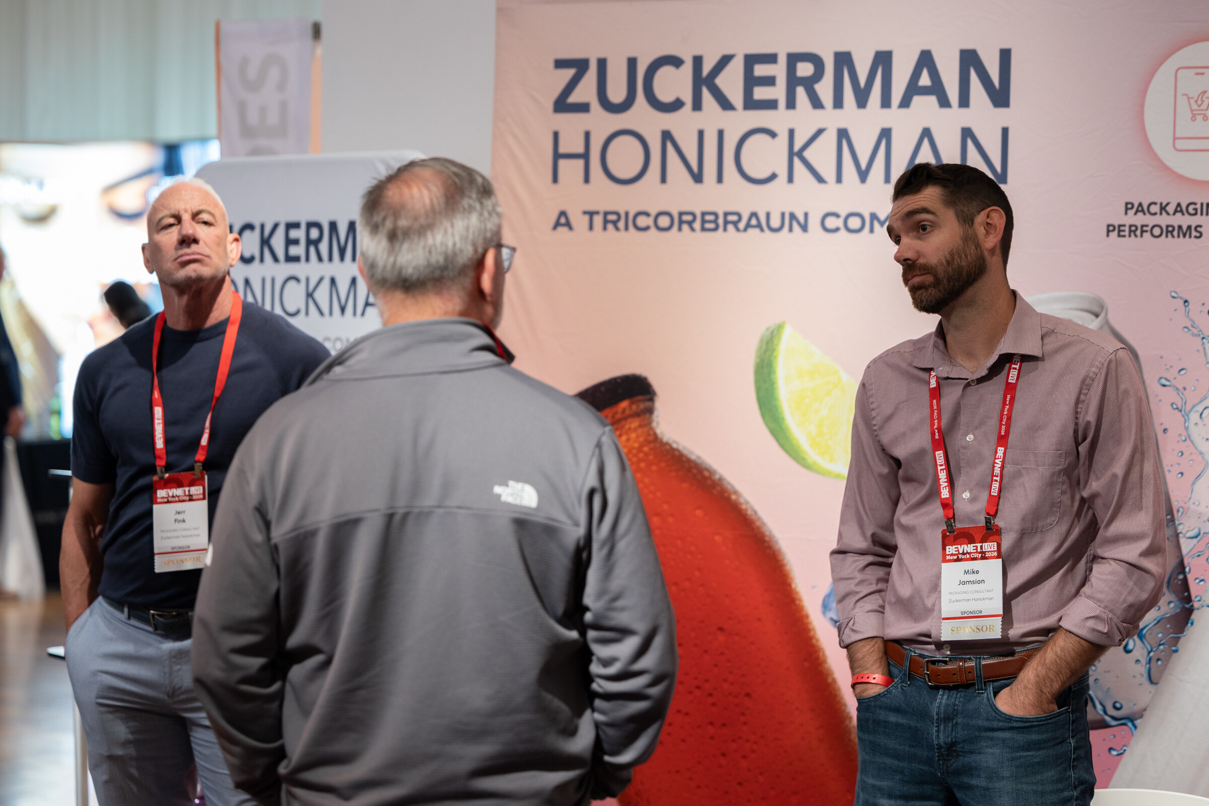 Three men talking in front of a Zuckerman Honickman, a TricorBraun company, booth banner.