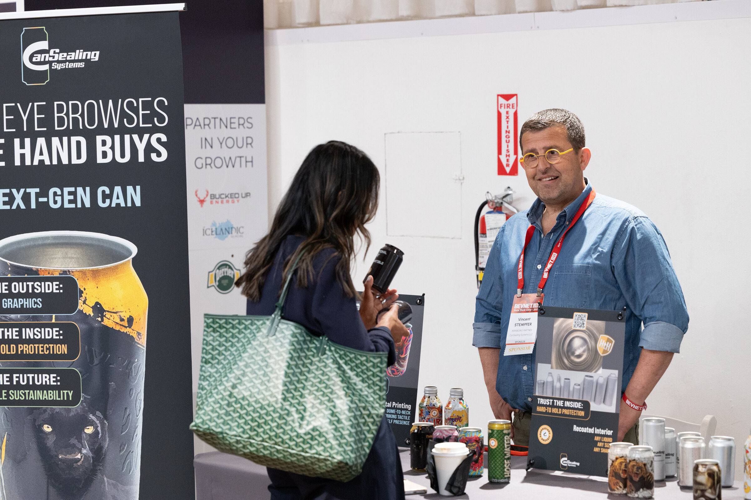 Woman holding a can talking with a smiling exhibitor in glasses at the CanSealing Systems booth, with a next-gen can banner.