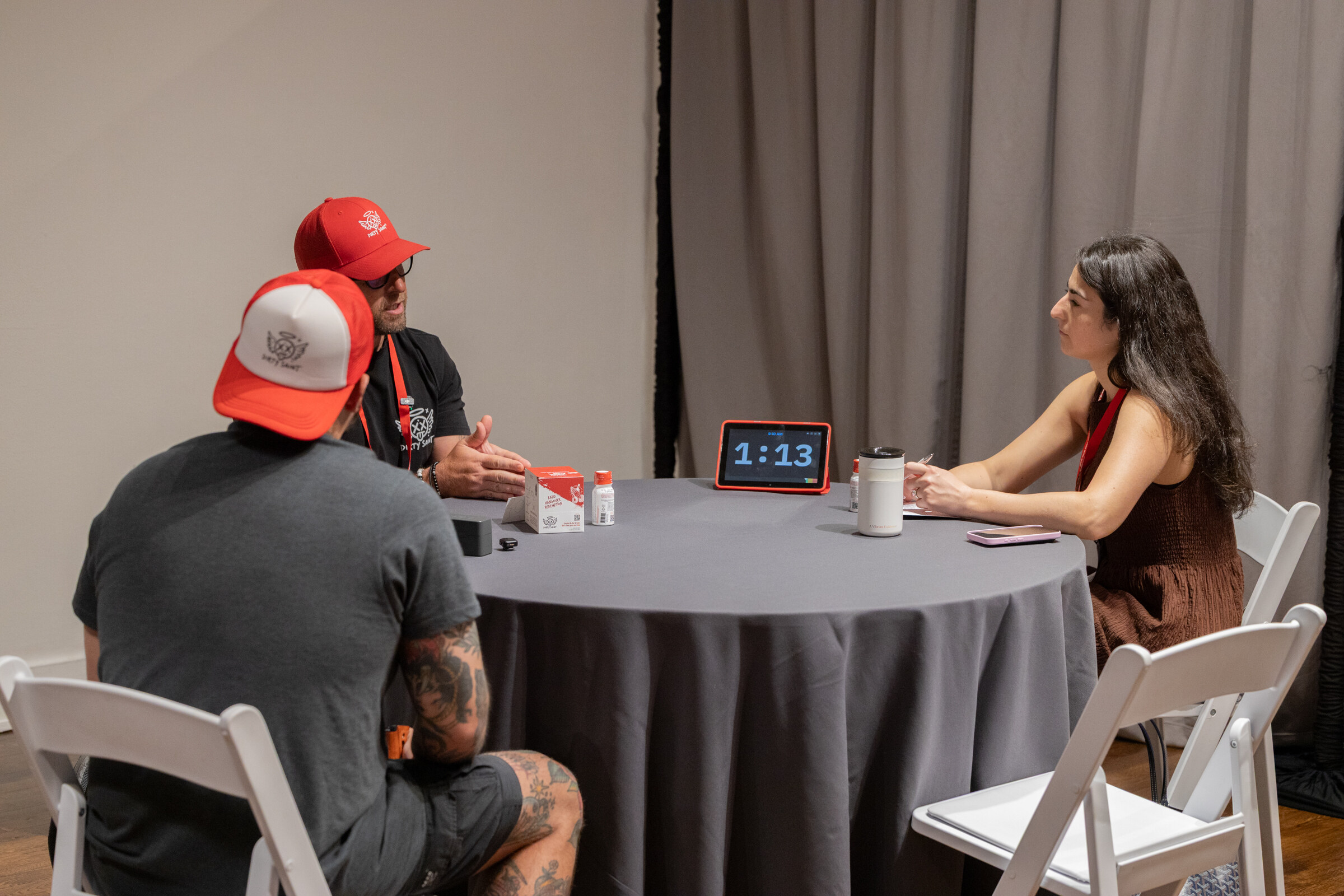 Two men in Dirty Saint caps meeting with a woman at a round table, a timer showing 1:13 between them.