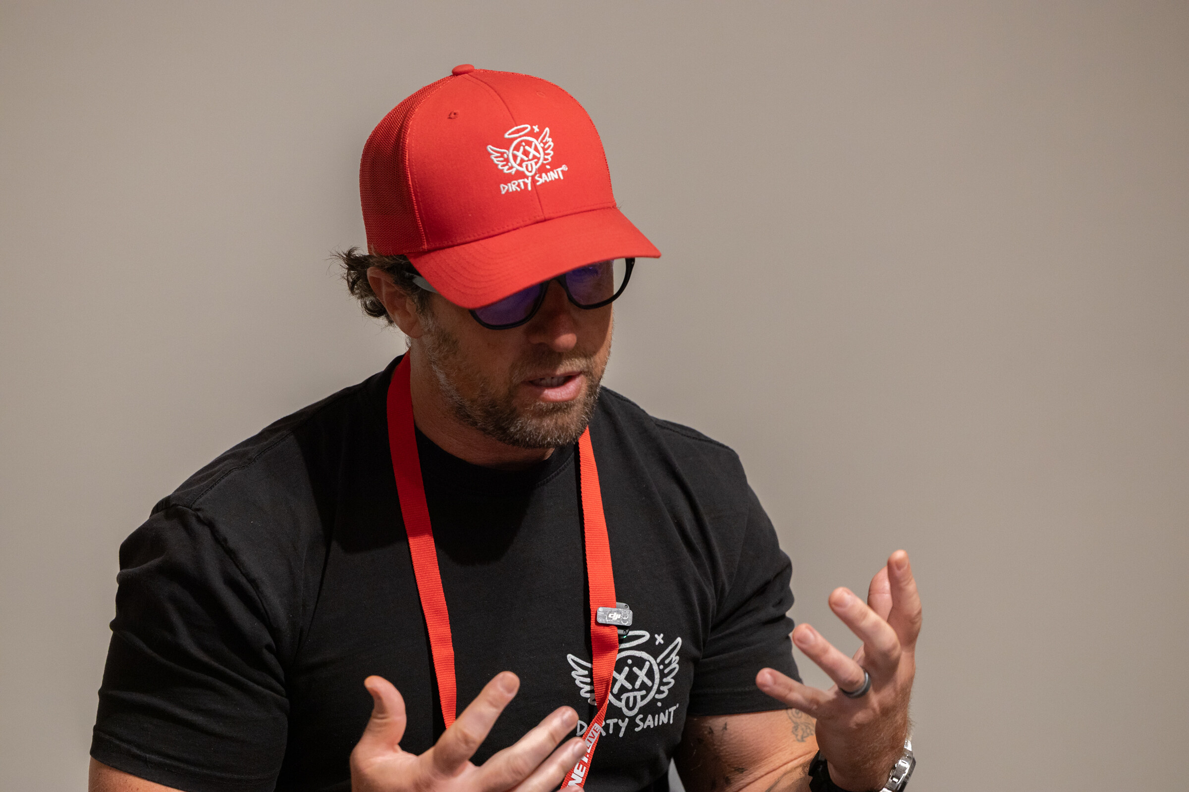 Man in a red Dirty Saint cap and sunglasses gesturing with both hands while speaking.