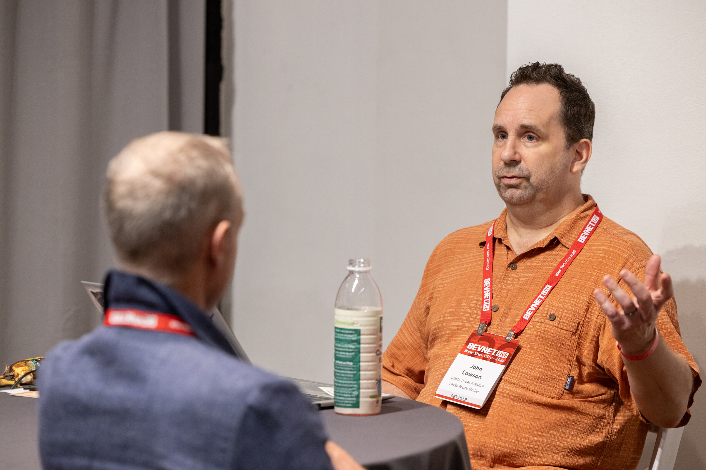 Man in an orange shirt with a BevNET Live lanyard gesturing as he talks with an attendee across a table.