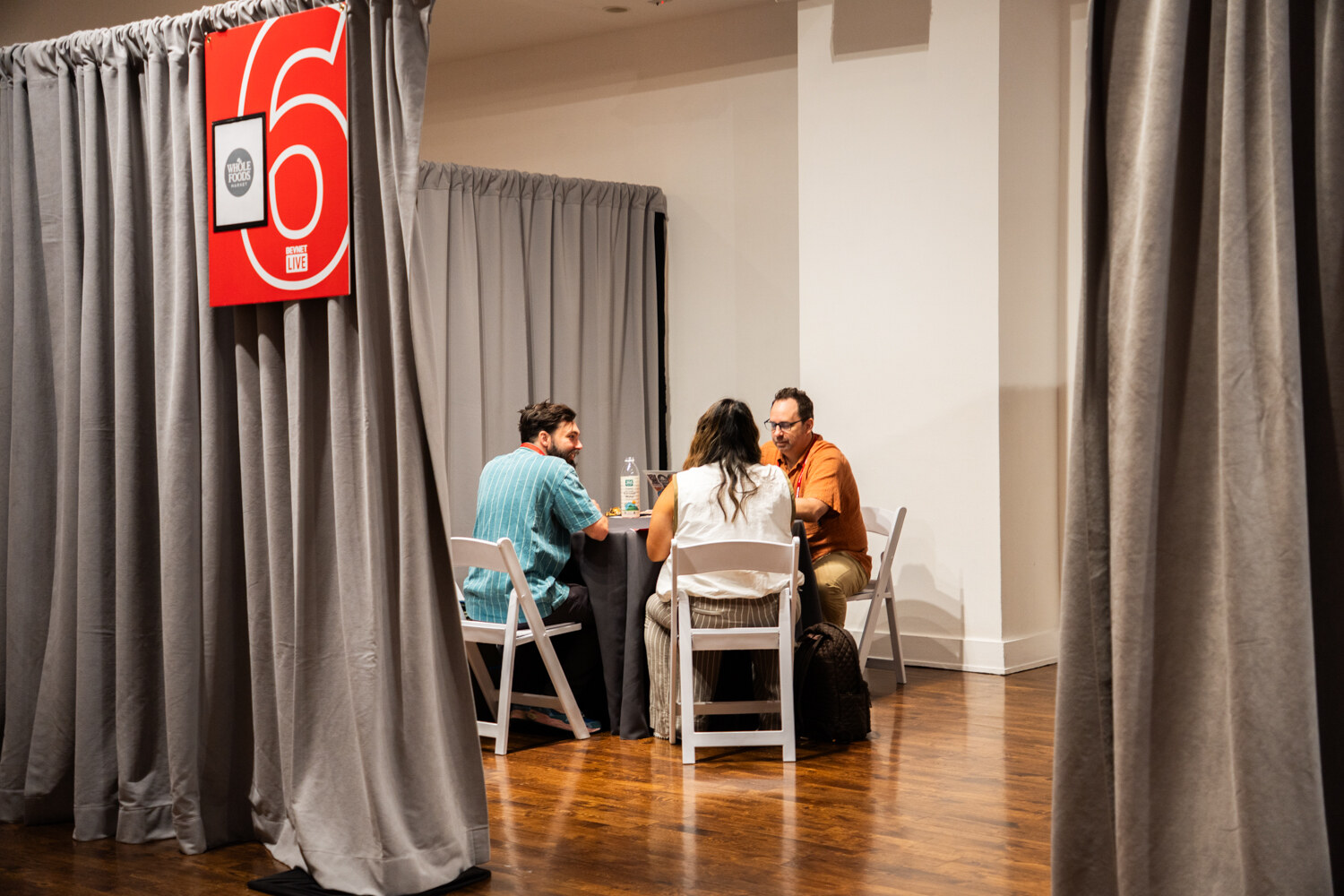View past a gray curtain marked with a large number 6 into a booth where three people meet at a round table.