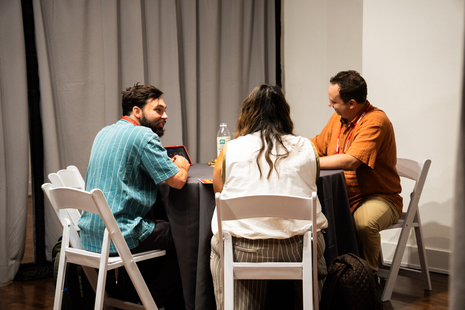 Three people at a round gray table in a curtained booth, a man in a teal shirt and a man in orange both smiling.