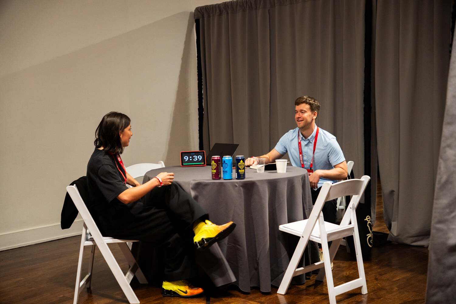 Woman in black with yellow shoes meeting a smiling man in a blue shirt at a round table with a timer and cans.