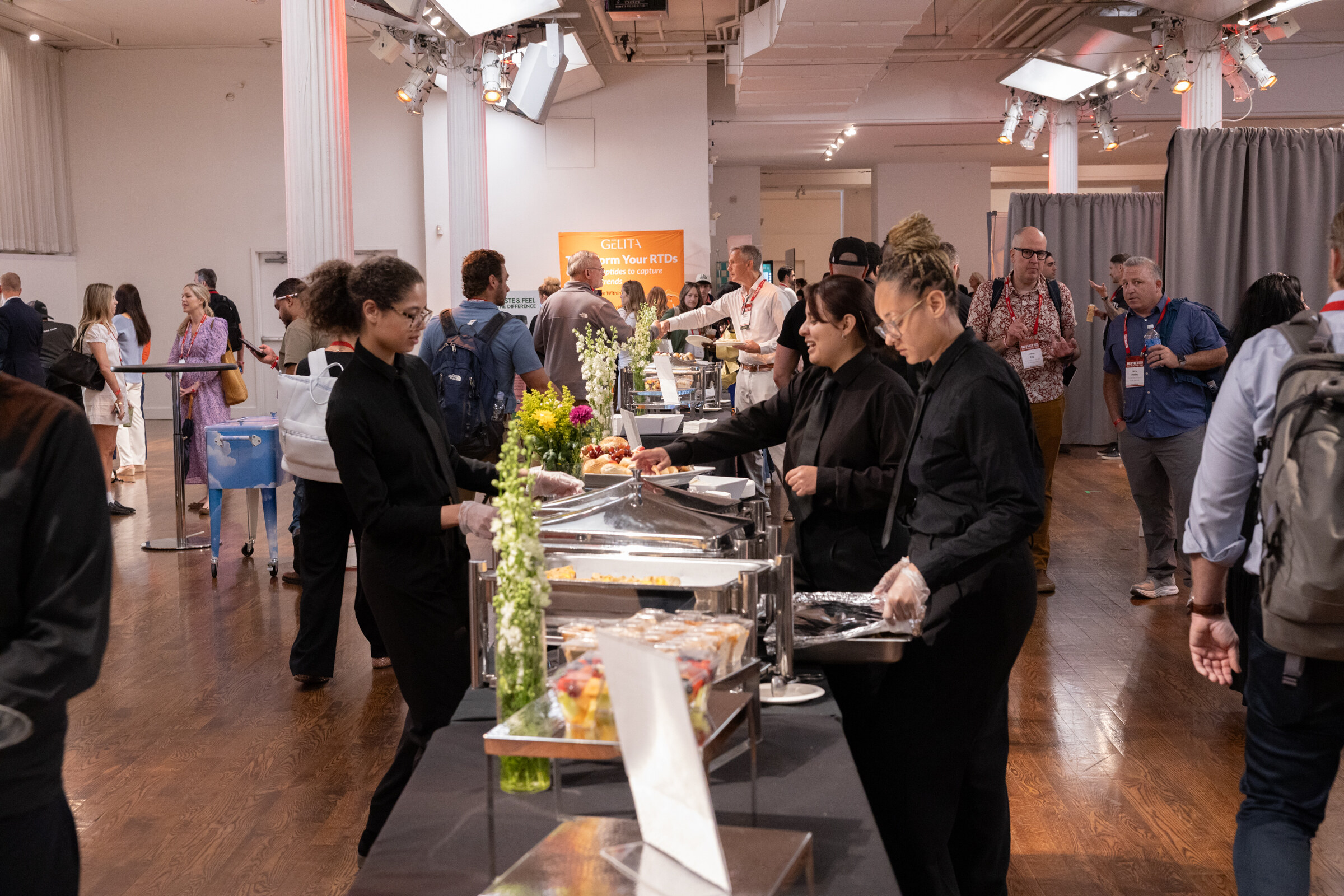 Catering staff in black serving from chafing dishes on a buffet line while attendees mingle across the hardwood floor.