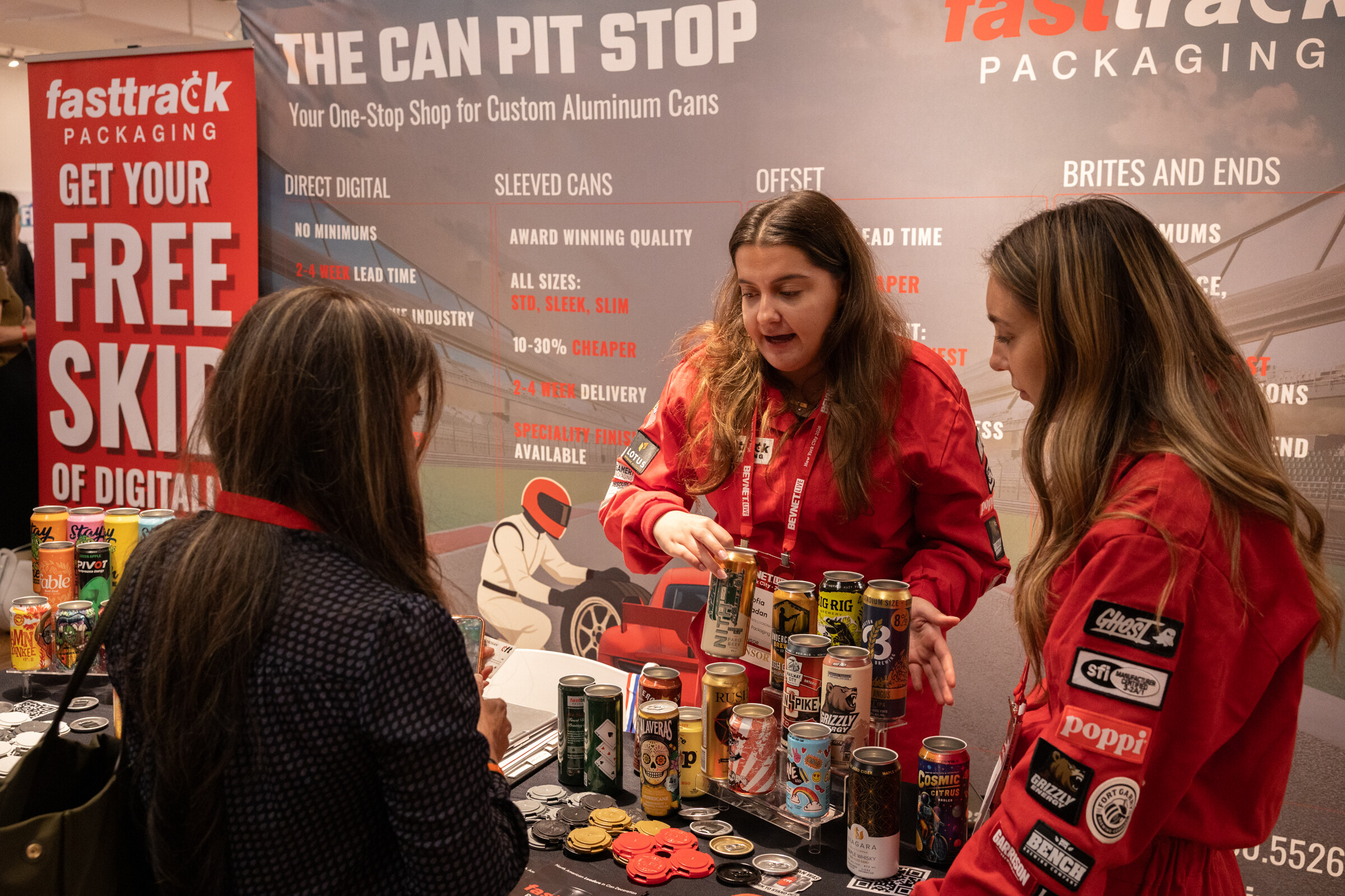 Two women in red racing jumpsuits showing printed cans to an attendee at the fasttrack Packaging Can Pit Stop booth.
