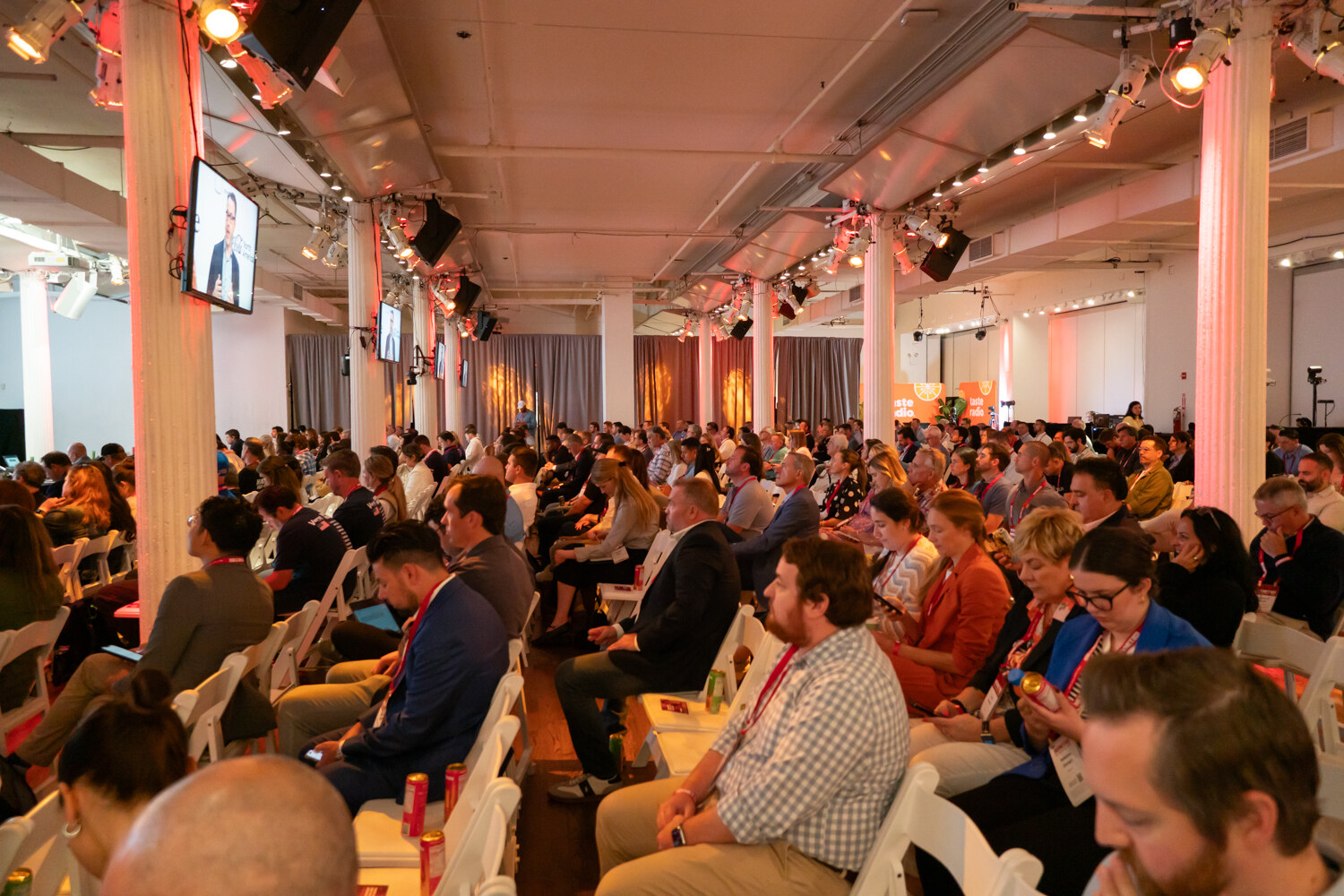 Wide view of a packed audience in rows of white chairs under columns and stage lights in a long event hall.