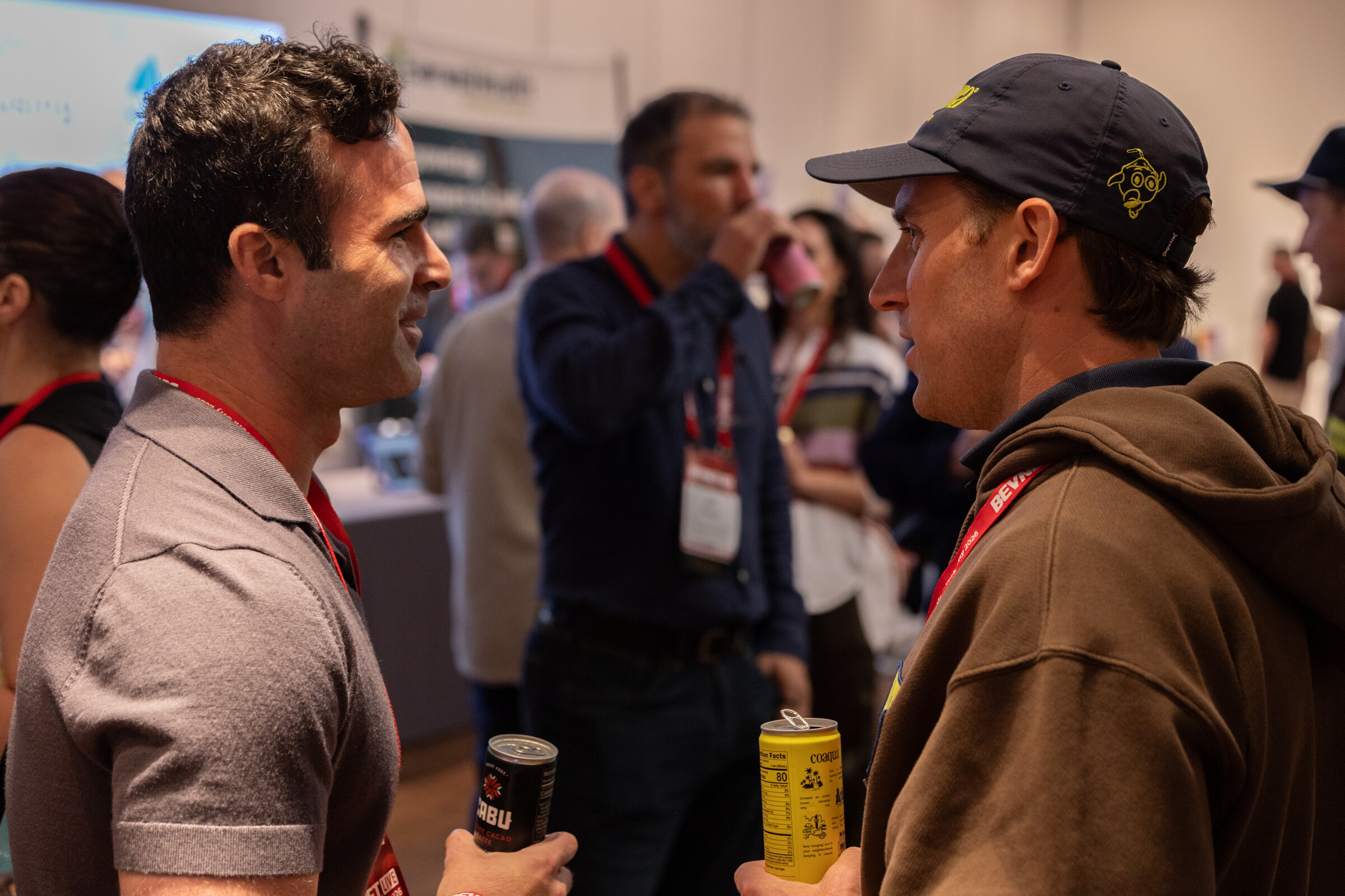 Two men in conversation holding beverage cans, one in a cap and brown hoodie.