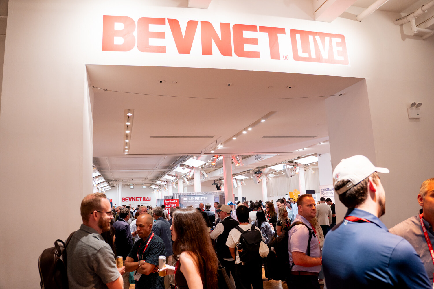 A crowd of attendees with lanyards gathered under and beyond a white archway topped by a large red BevNET Live sign.