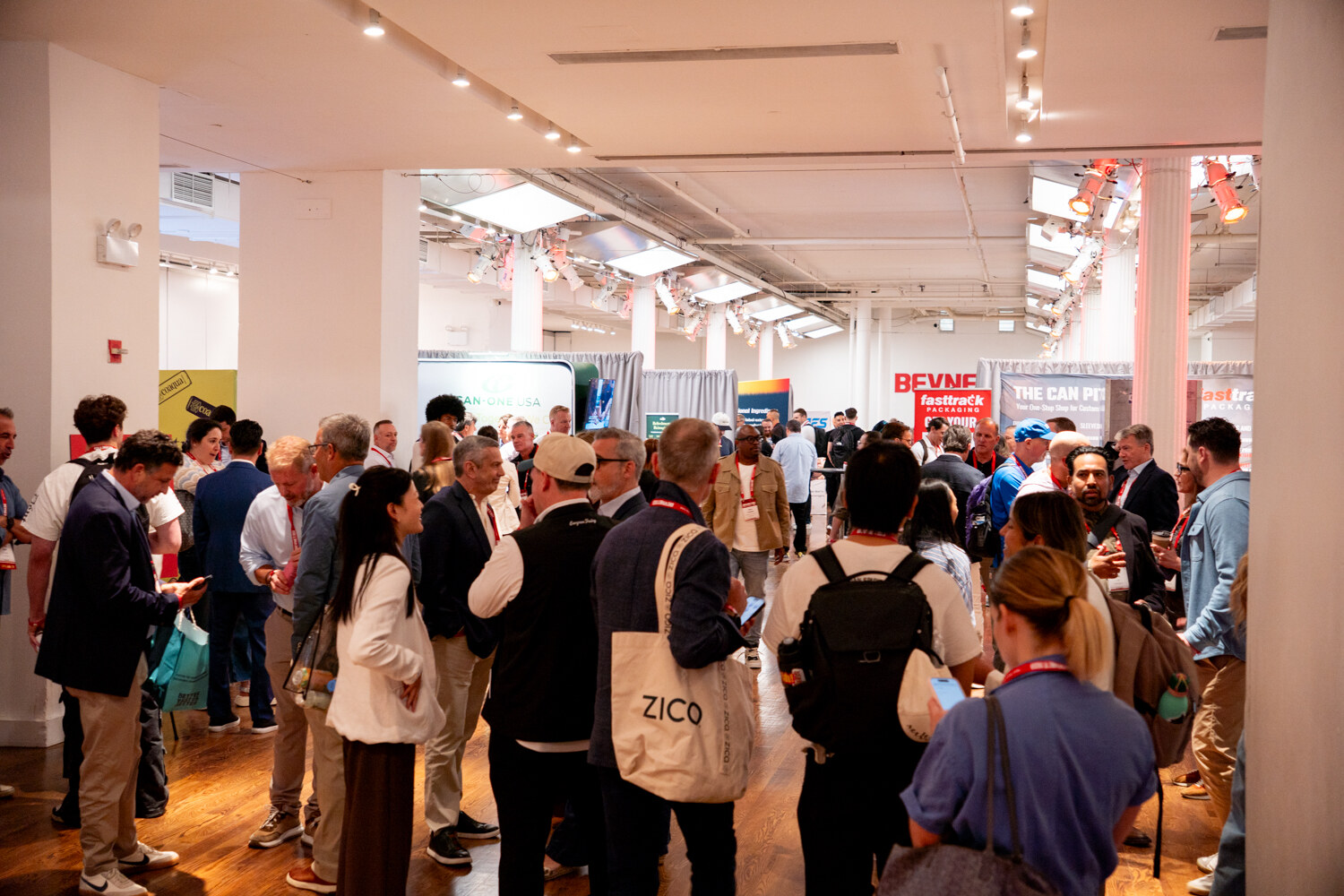 A dense crowd of attendees talking among sponsor booths on a hardwood floor, with a BevNET Live sign in the distance.