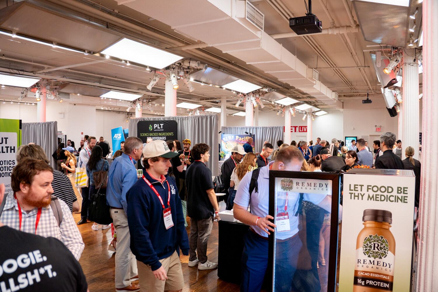 Attendees walking past sponsor booths, including a display for Remedy Organics protein drinks reading Let Food Be Thy Medicine.
