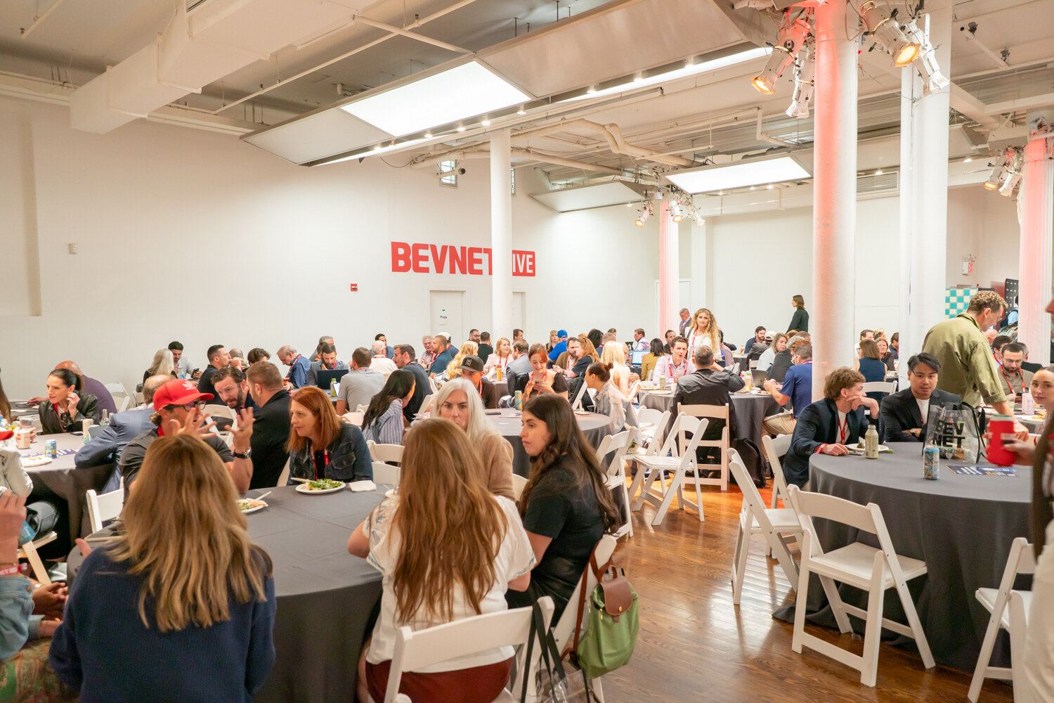 Dozens of attendees eating lunch at round gray tables in a white room with a red BevNET Live sign on the wall.