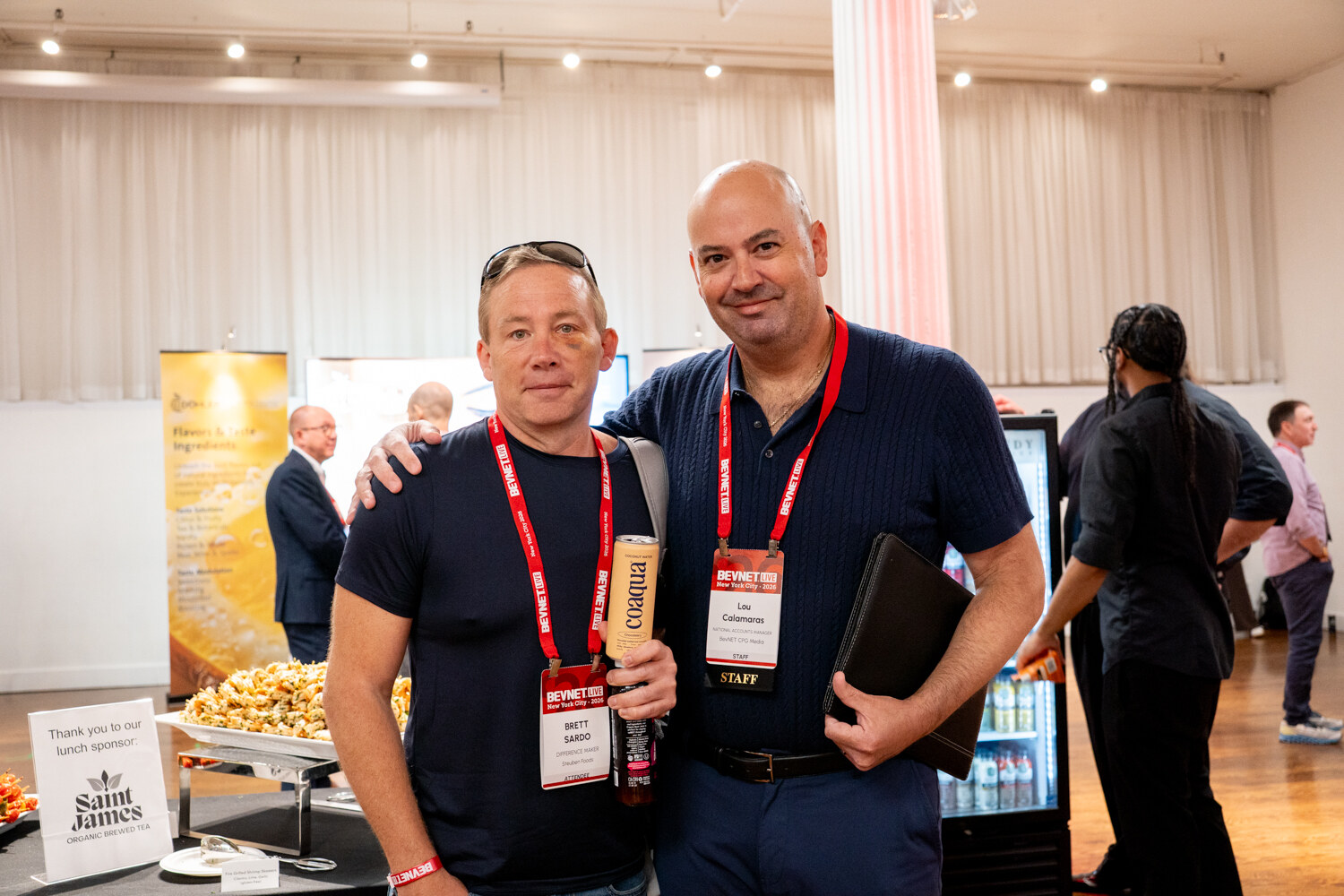 Two men in navy shirts with BevNET Live lanyards posing arm in arm, one holding a coaqua can, near a lunch sponsor sign.