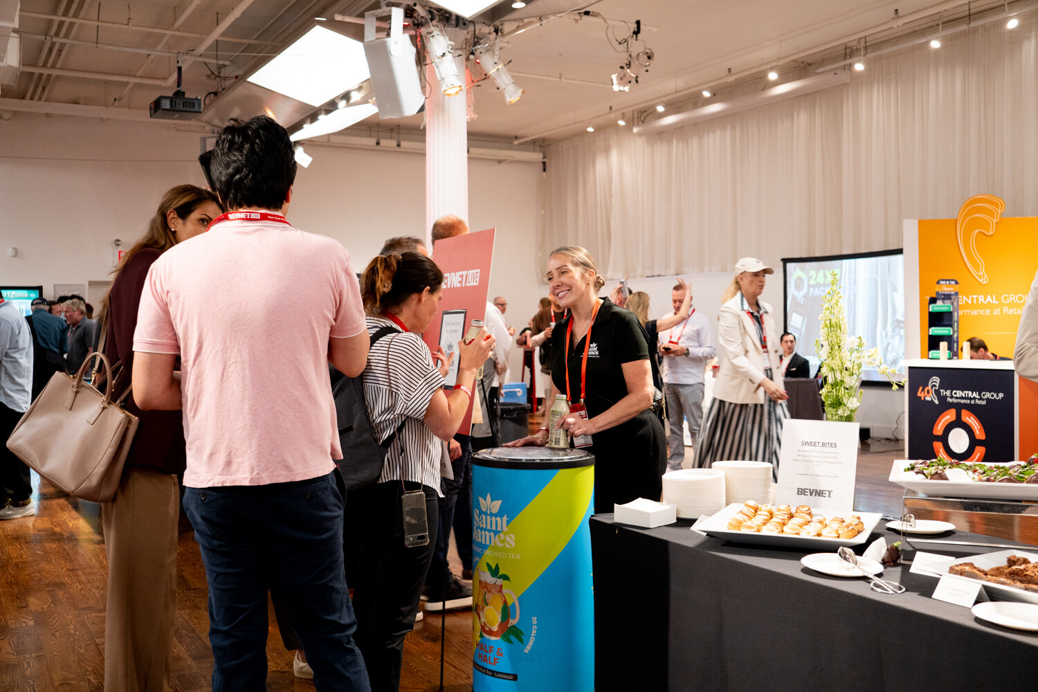 Woman in a black shirt smiling at attendees at a food station next to a blue and yellow Saint James tea cooler.
