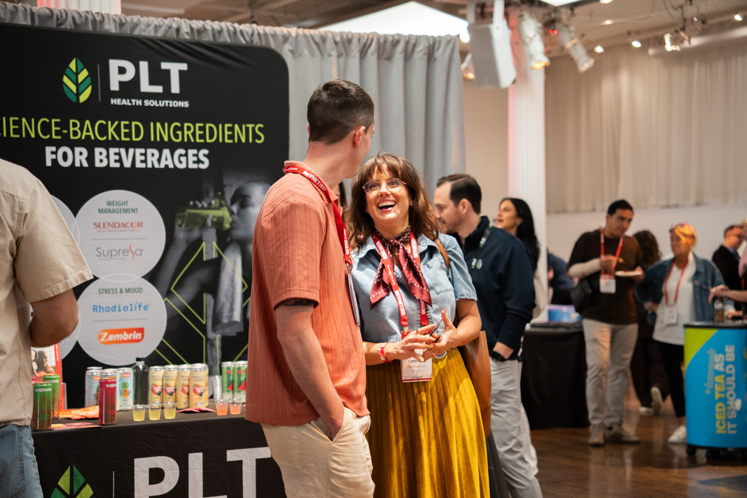 Smiling woman in a yellow skirt talking with a man in a salmon shirt beside a PLT Health Solutions booth banner.