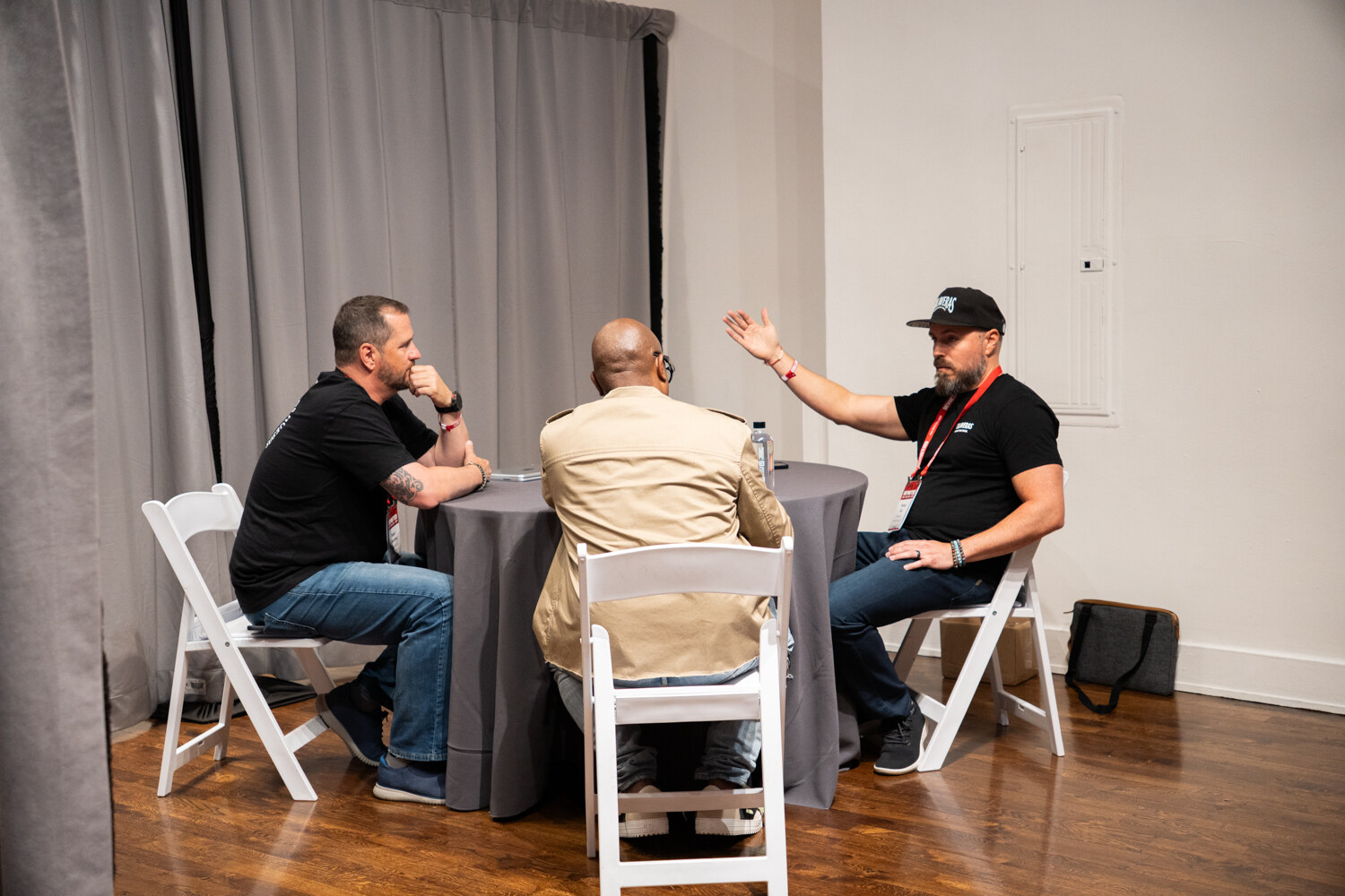 Man in a black cap gesturing while two others listen at a round table in a curtained booth.