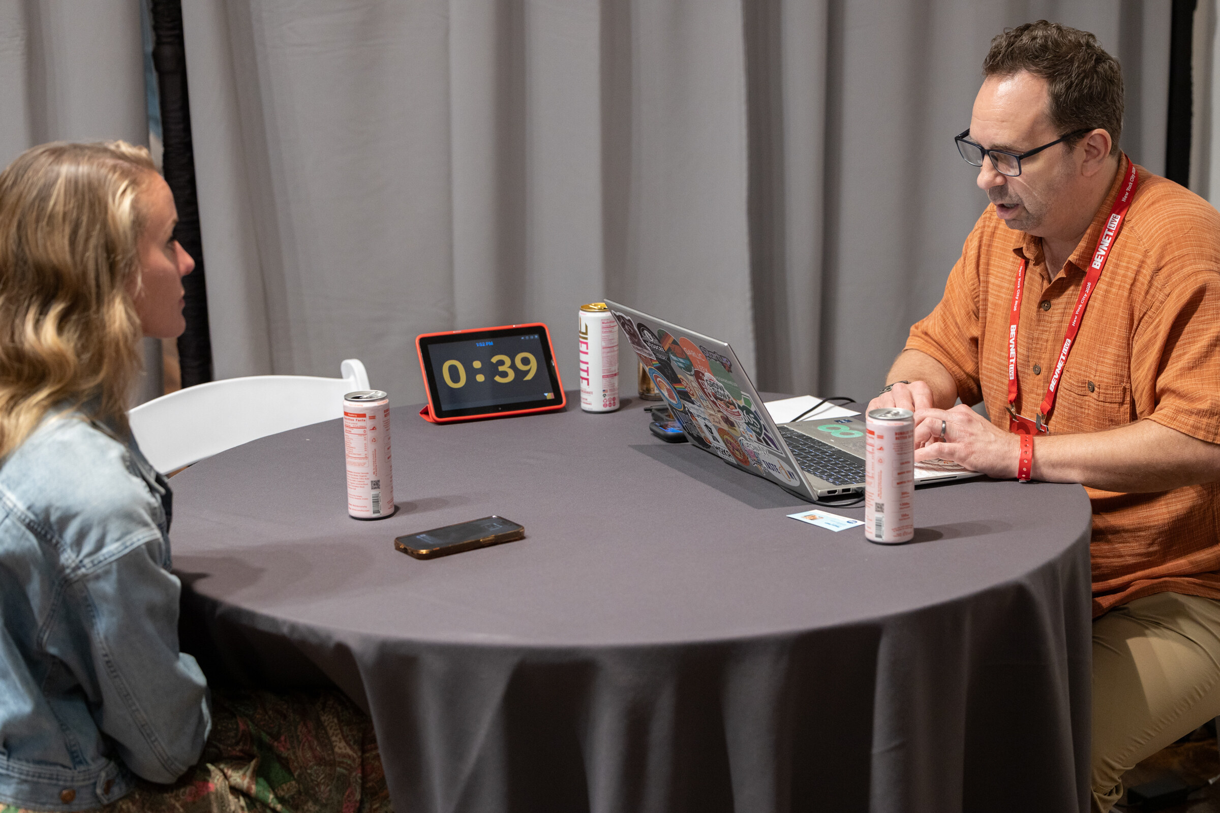 A host typing on a laptop across from a woman, with cans and a timer reading 0:39 on the table.