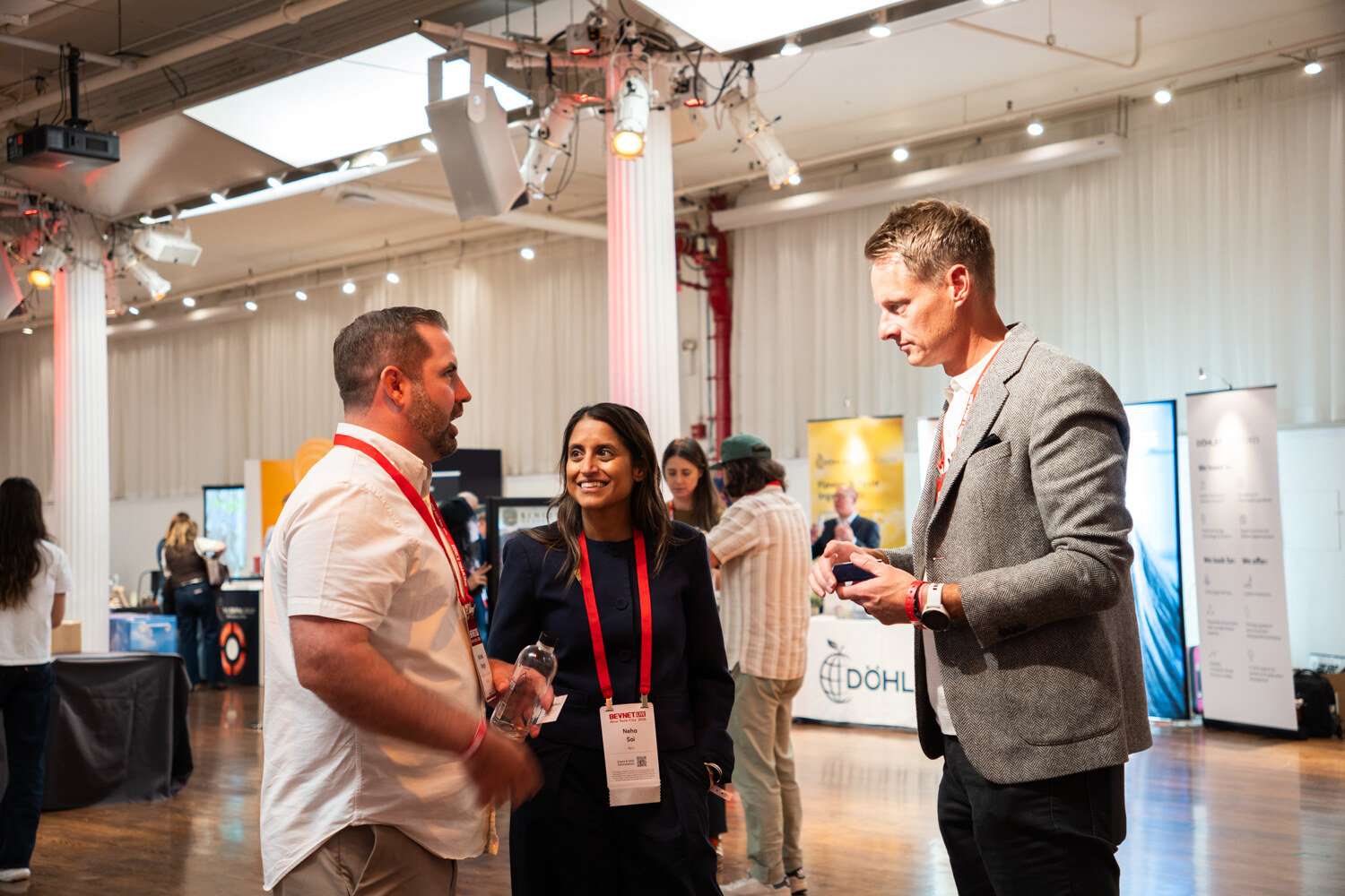 Three attendees in conversation on the expo floor, one man holding a phone, with booths behind.