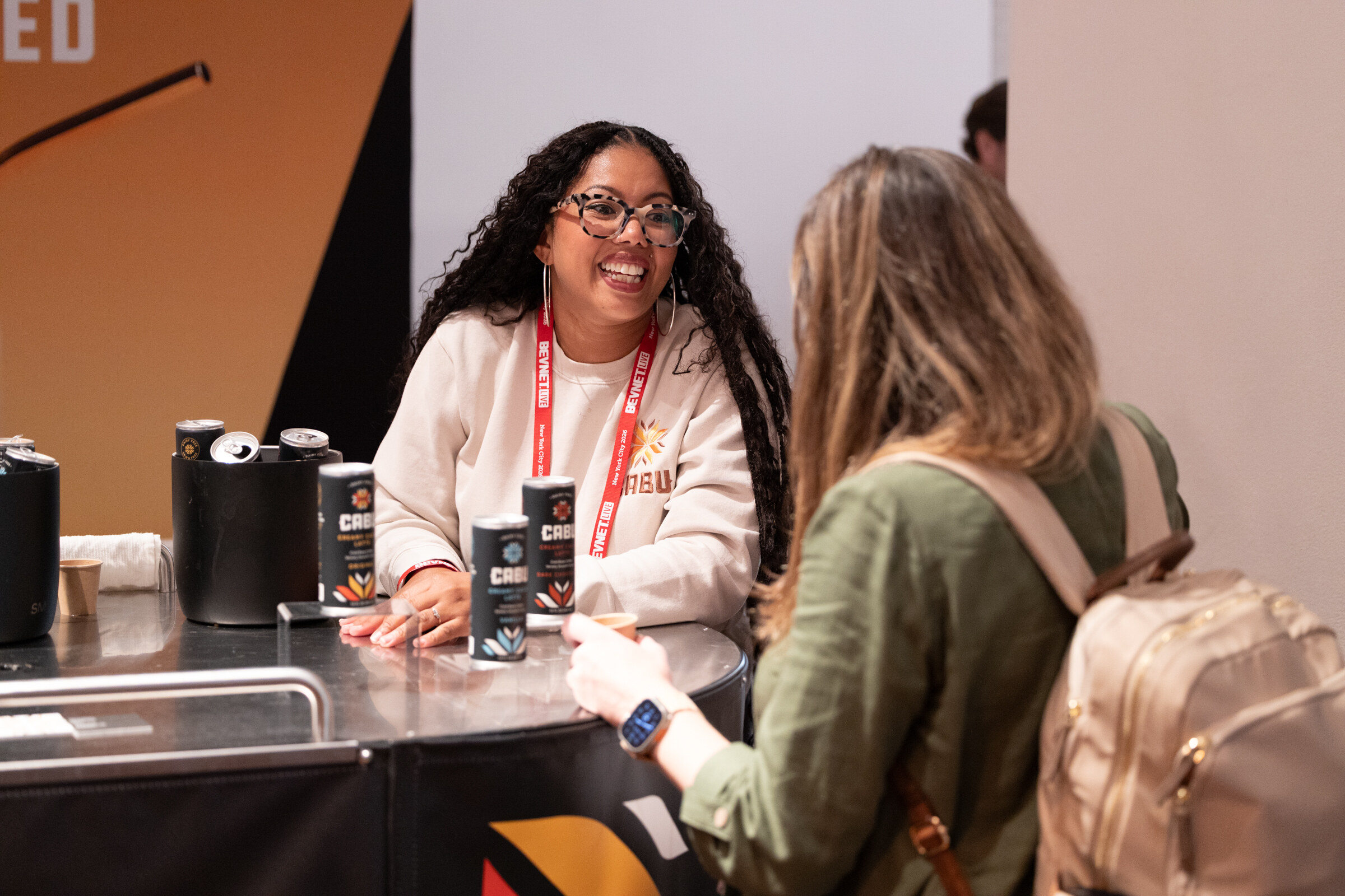 Woman in glasses smiling behind a counter with cans of CABU while talking with an attendee.