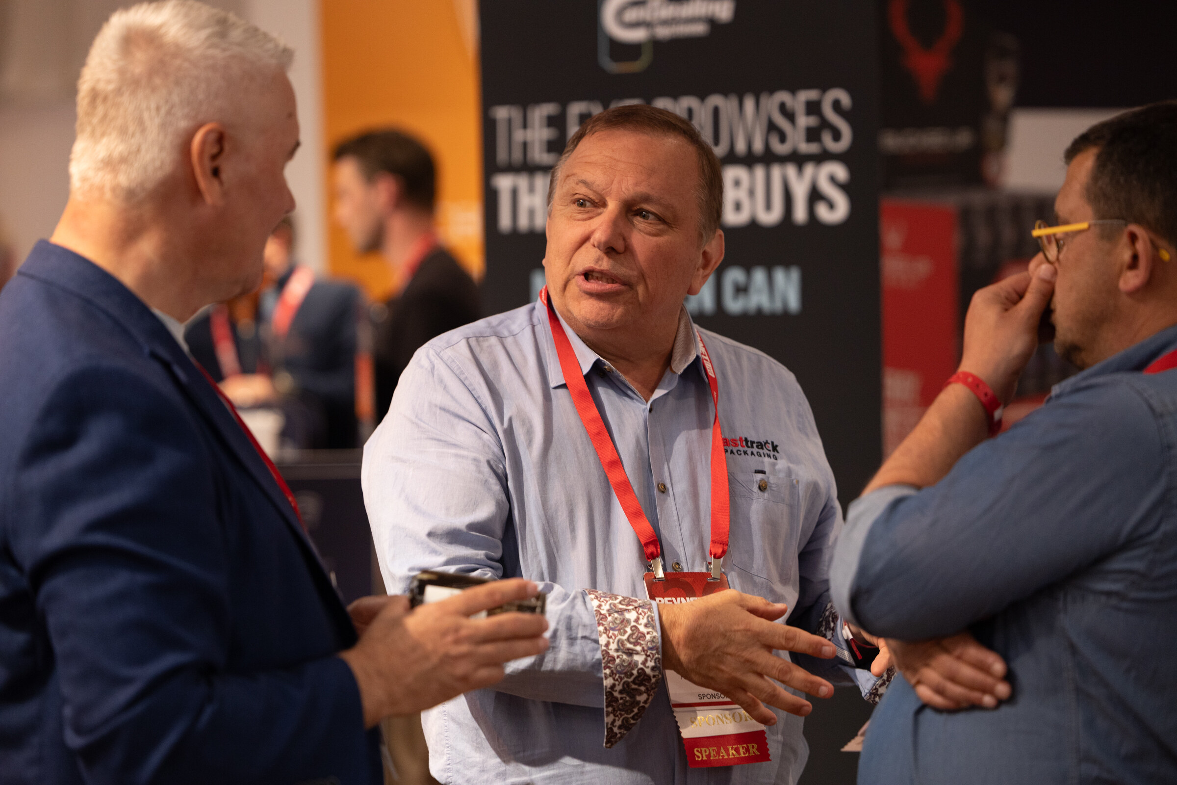 Man with a speaker badge gesturing as he talks with two attendees in front of a sponsor banner.
