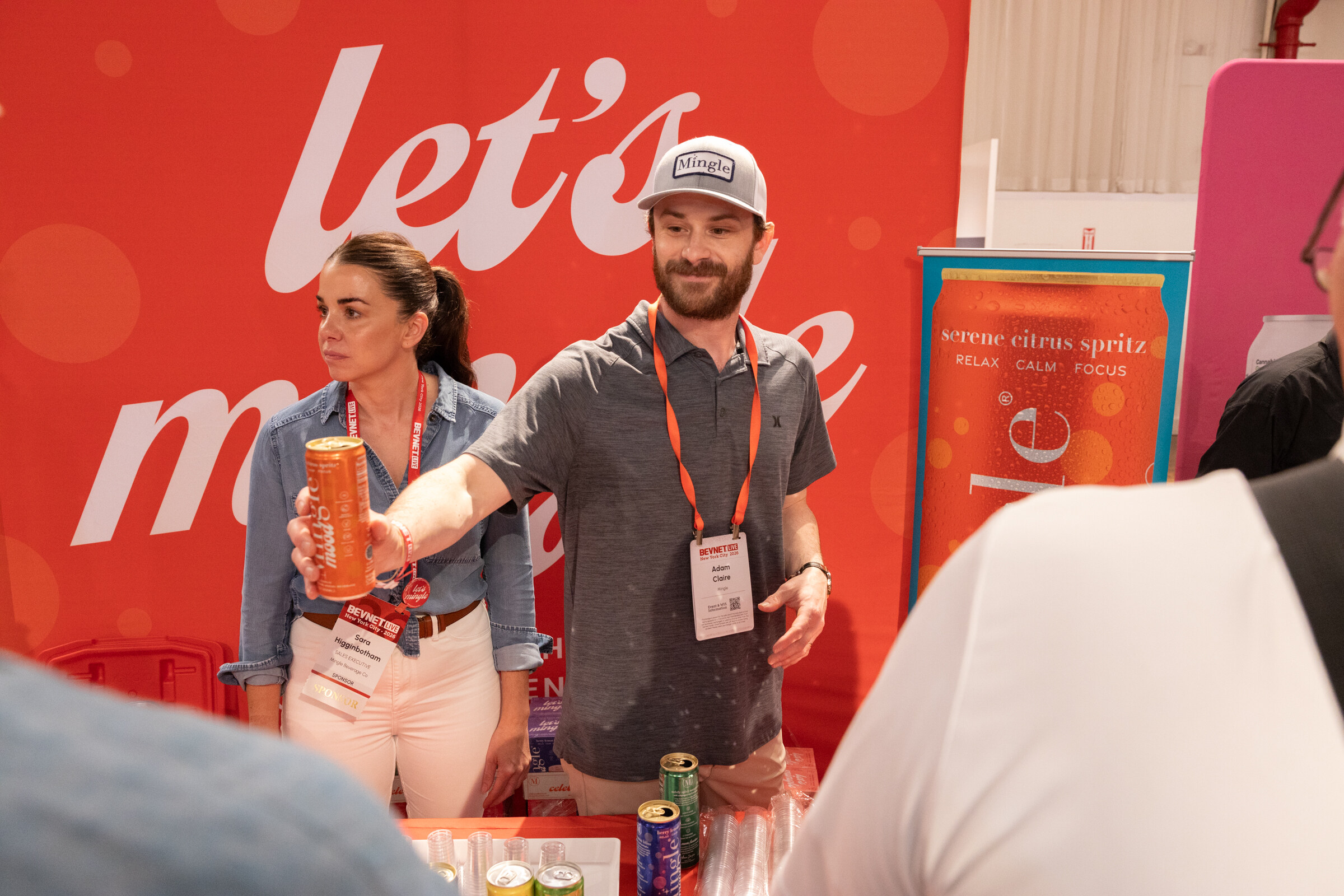 Man and woman behind the Mingle booth, the man handing a can across the table in front of a red let’s mingle backdrop.