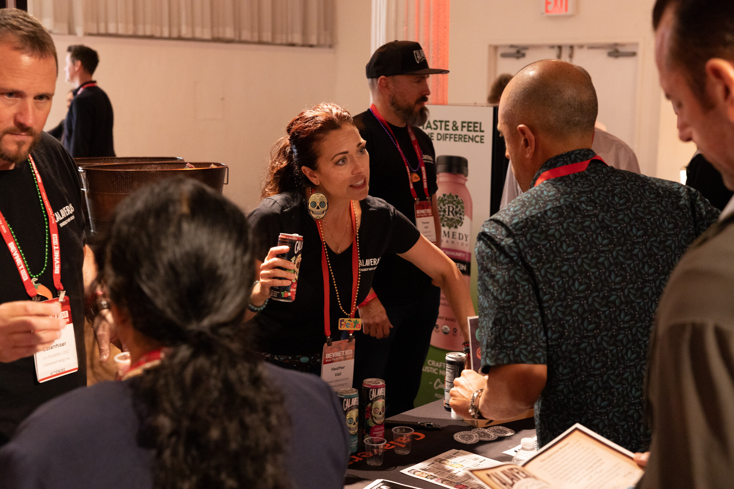 Woman in a black Calaveras shirt holding a can and talking with attendees across a sampling table.