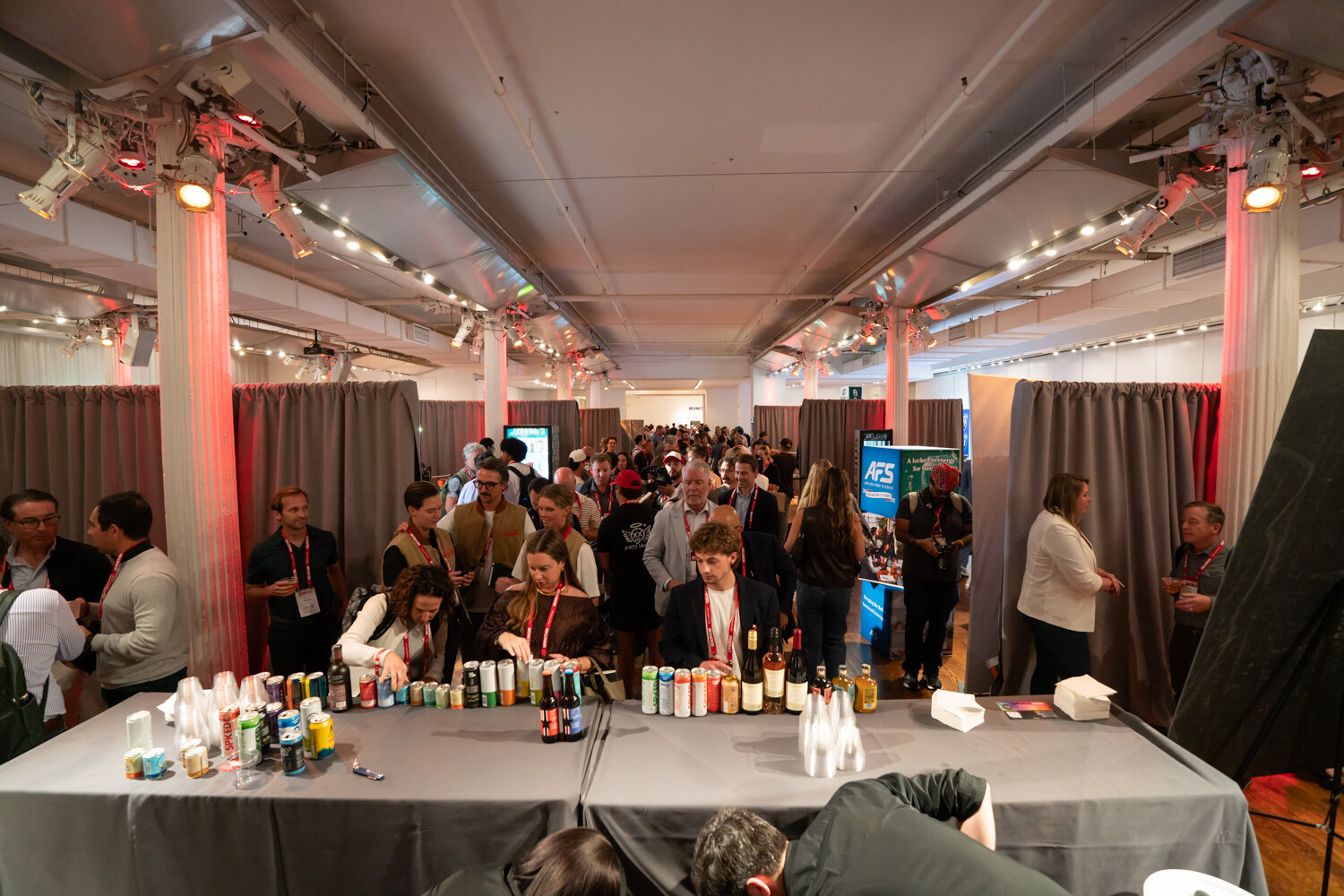 Wide view down a long gray tasting table lined with bottles and cans as a crowd of attendees fills the hall.