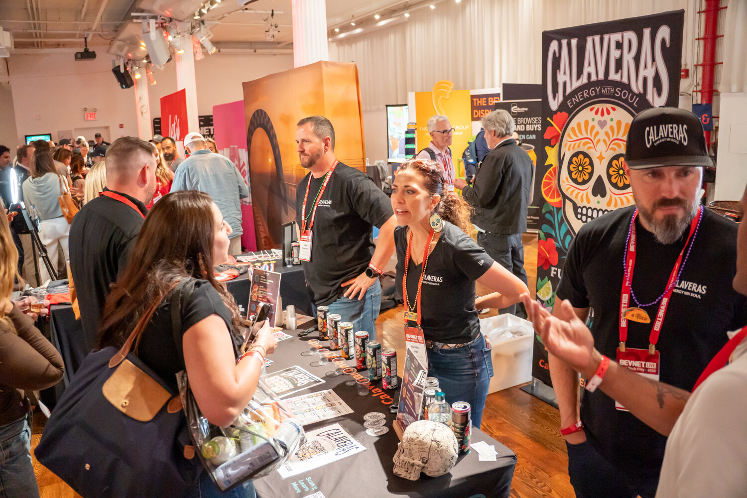 Calaveras Energy staff in black shirts talking with attendees at a table of cans in front of a sugar skull banner.
