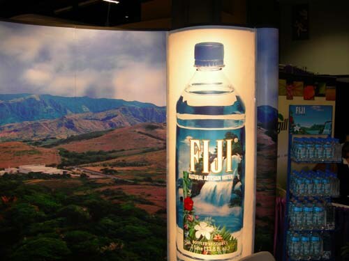 A lit display panel with a large FIJI Natural Artesian Water bottle in front of a landscape mural of green hills; a shelf of FIJI water bottles stands at right.