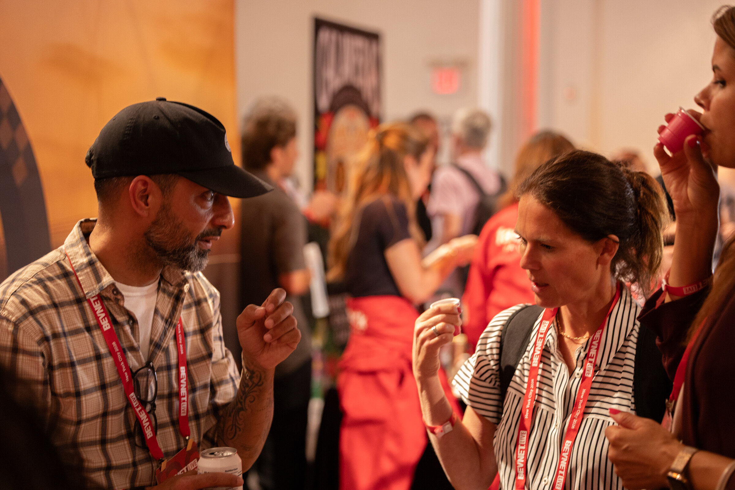 Man in a cap and plaid shirt talking with a woman sipping a sample in a crowded room.