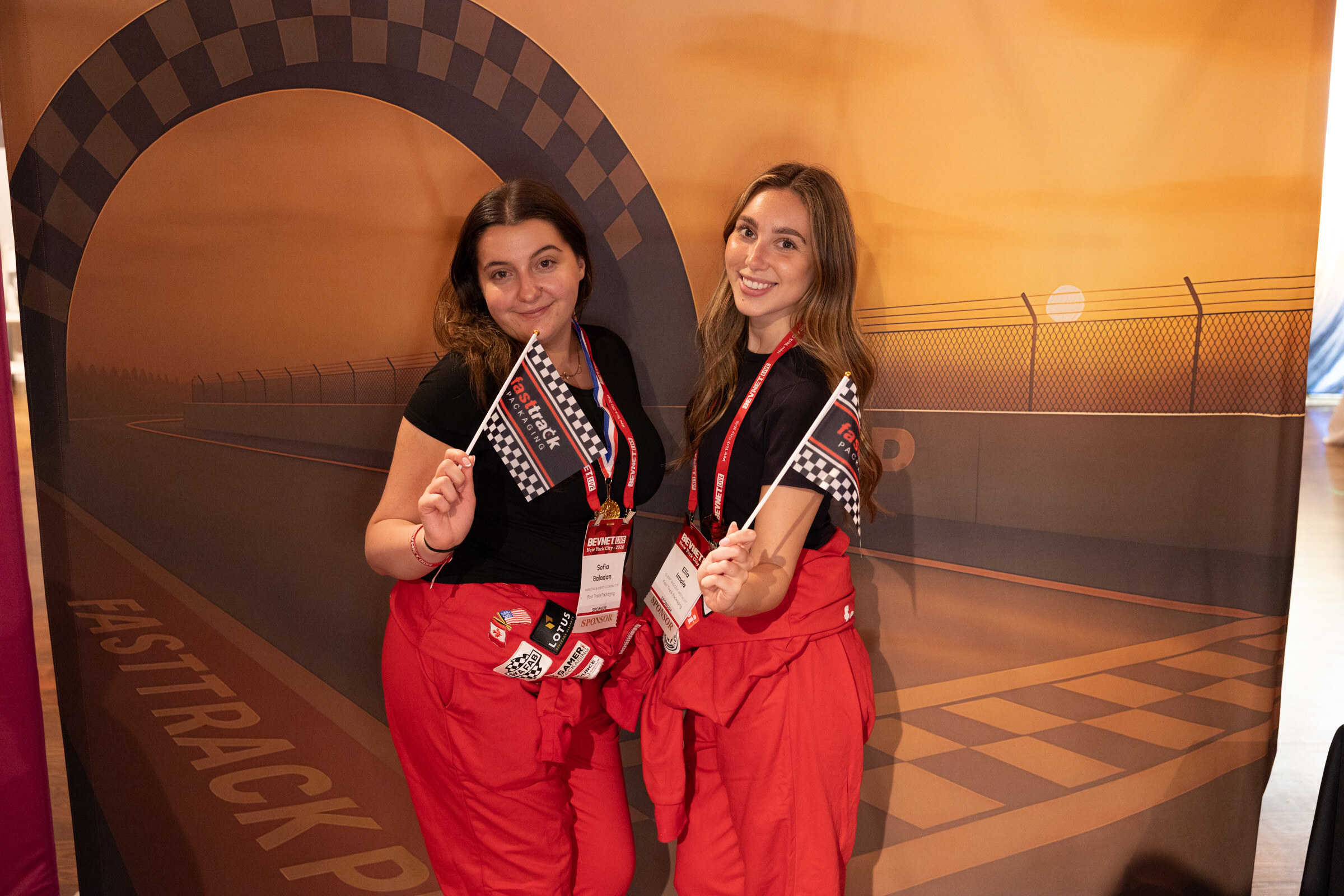 Two smiling women in red racing jumpsuits holding checkered flags in front of a racetrack backdrop.