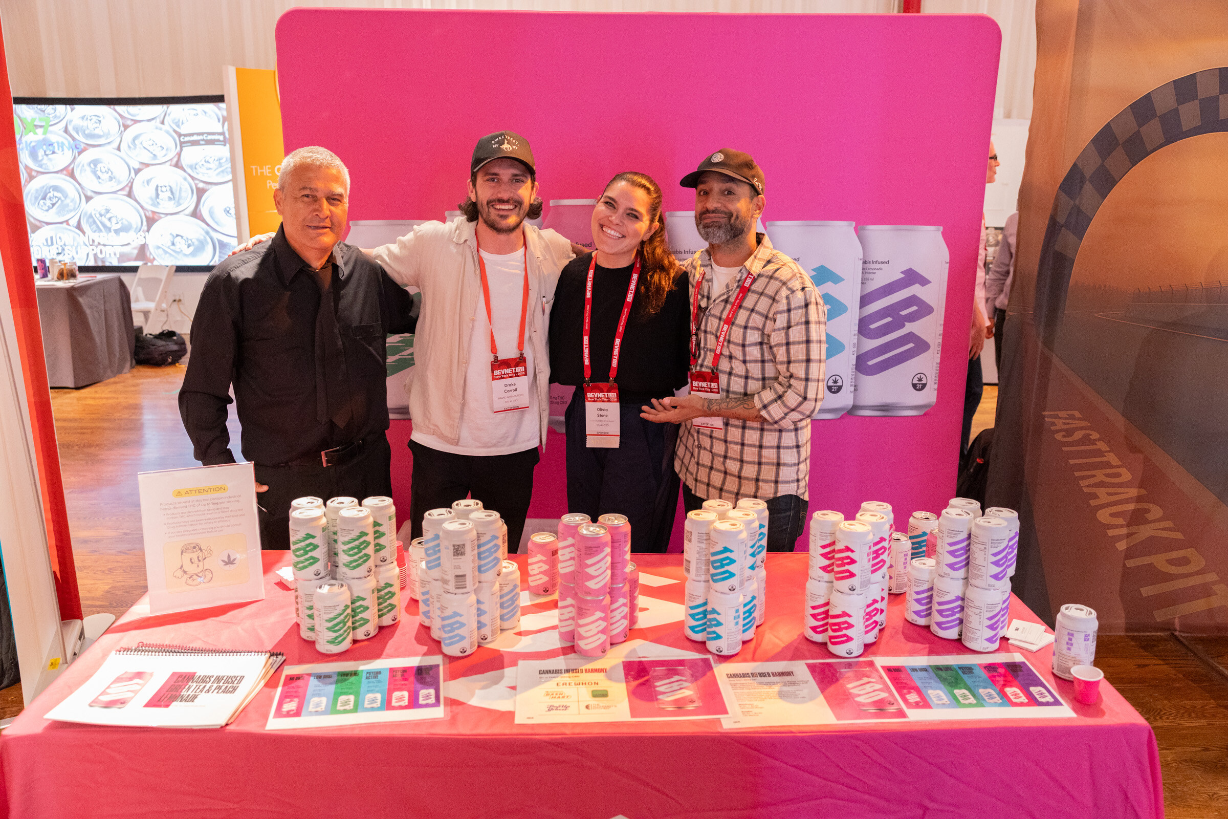 Four people posing behind a pink table stacked with cans in front of a pink and white booth backdrop.